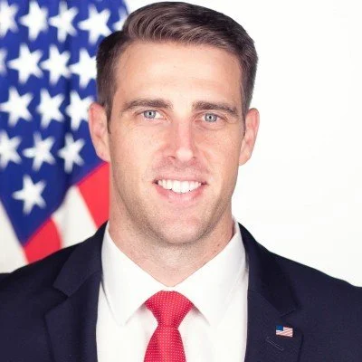Smiling young man standing outdoors in front of a red brick building with trees around, wearing a light-colored, patterned short-sleeve button-up shirt.