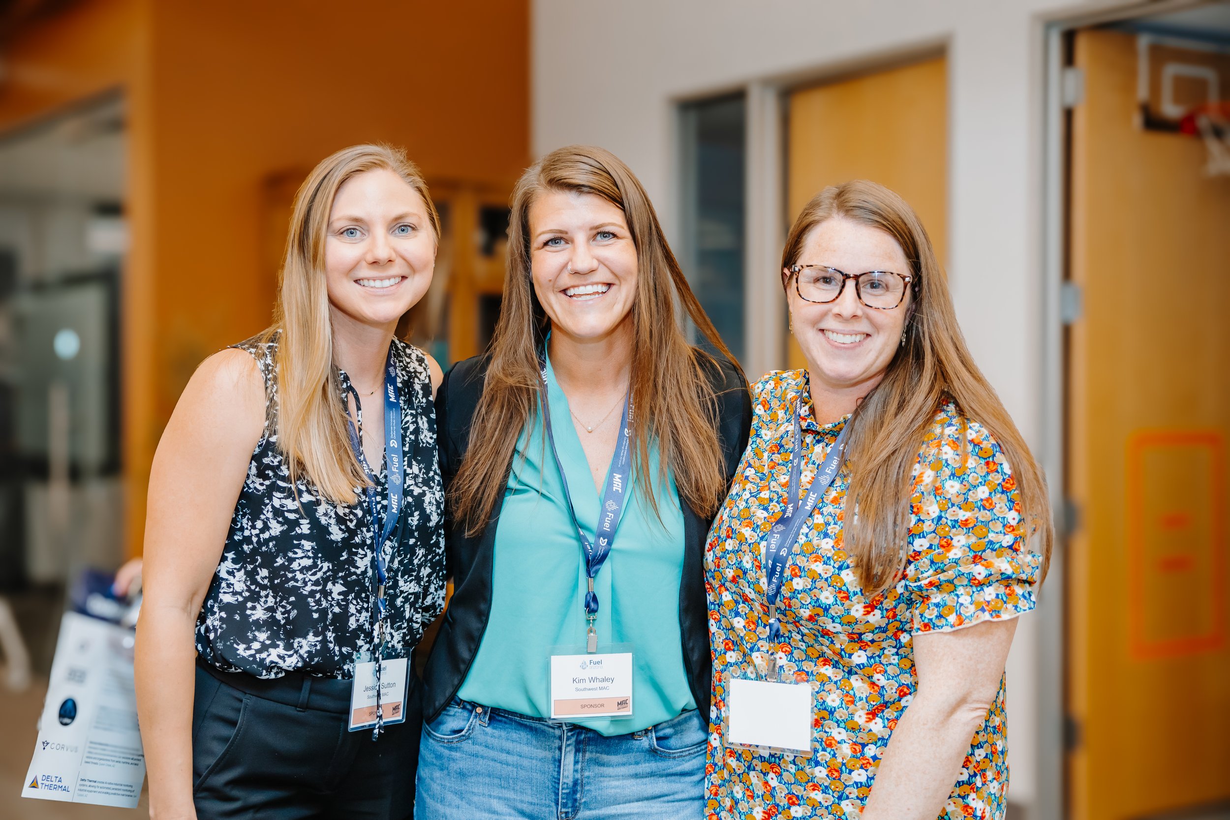 Three women smiling at a conference, wearing badges and lanyards, standing indoors with a yellow wall and windows in the background.