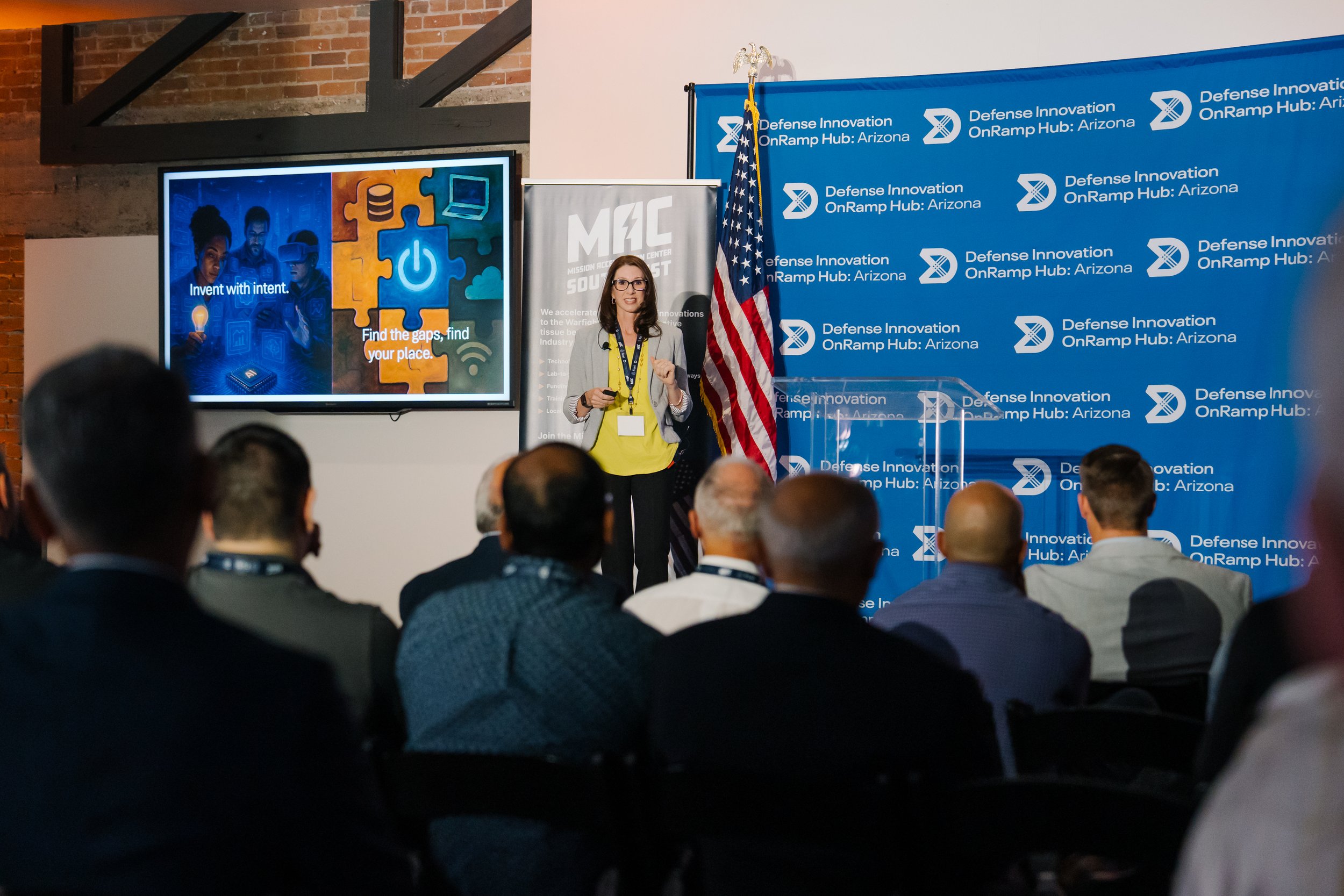 A woman giving a presentation at a conference with a blue backdrop showing the logo and name 'Defense Innovation OnRamp Hub: Arizona'. There is a large screen displaying a graphic with the text 'Invent with intent' and 'Find the gaps, find your place