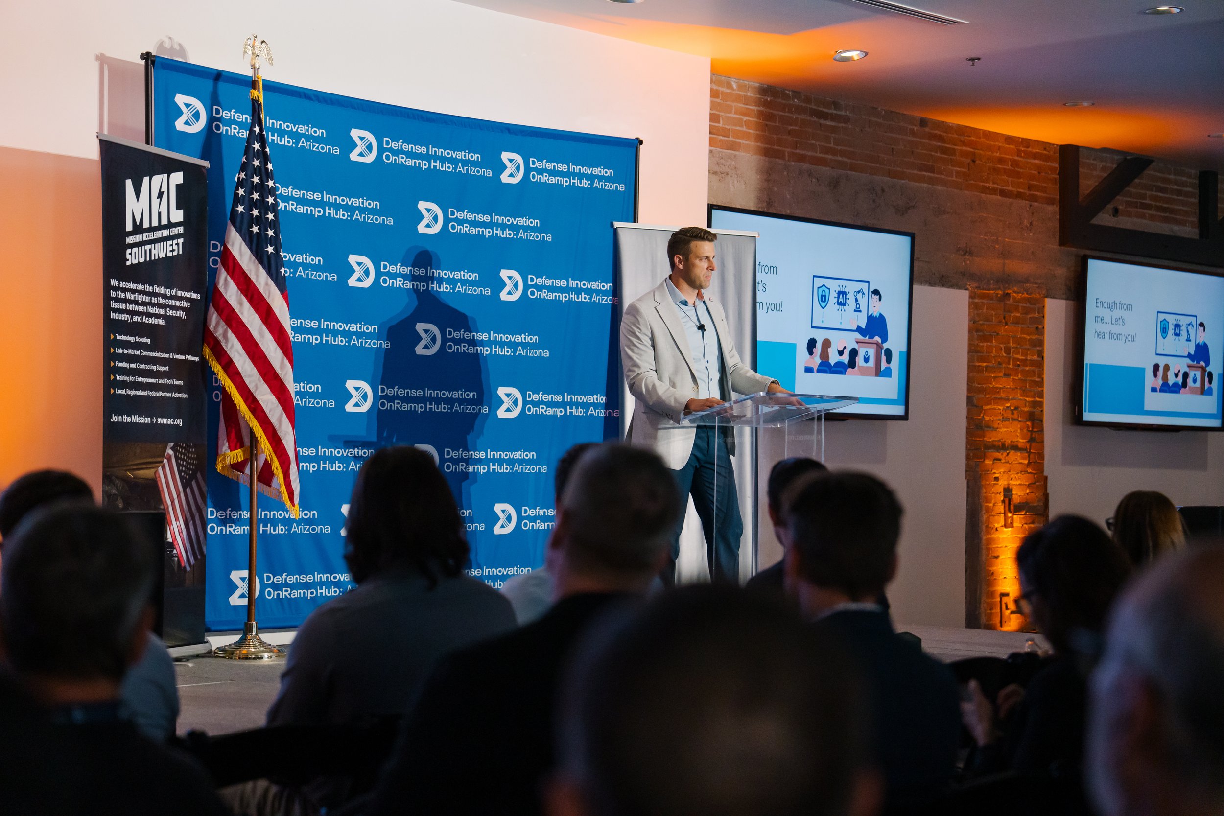 A man in a light-colored blazer giving a presentation at a conference with a blue backdrop that says "Defense Innovation OnRamp Hub: Arizona". There are two large screens displaying slides and an audience watching.