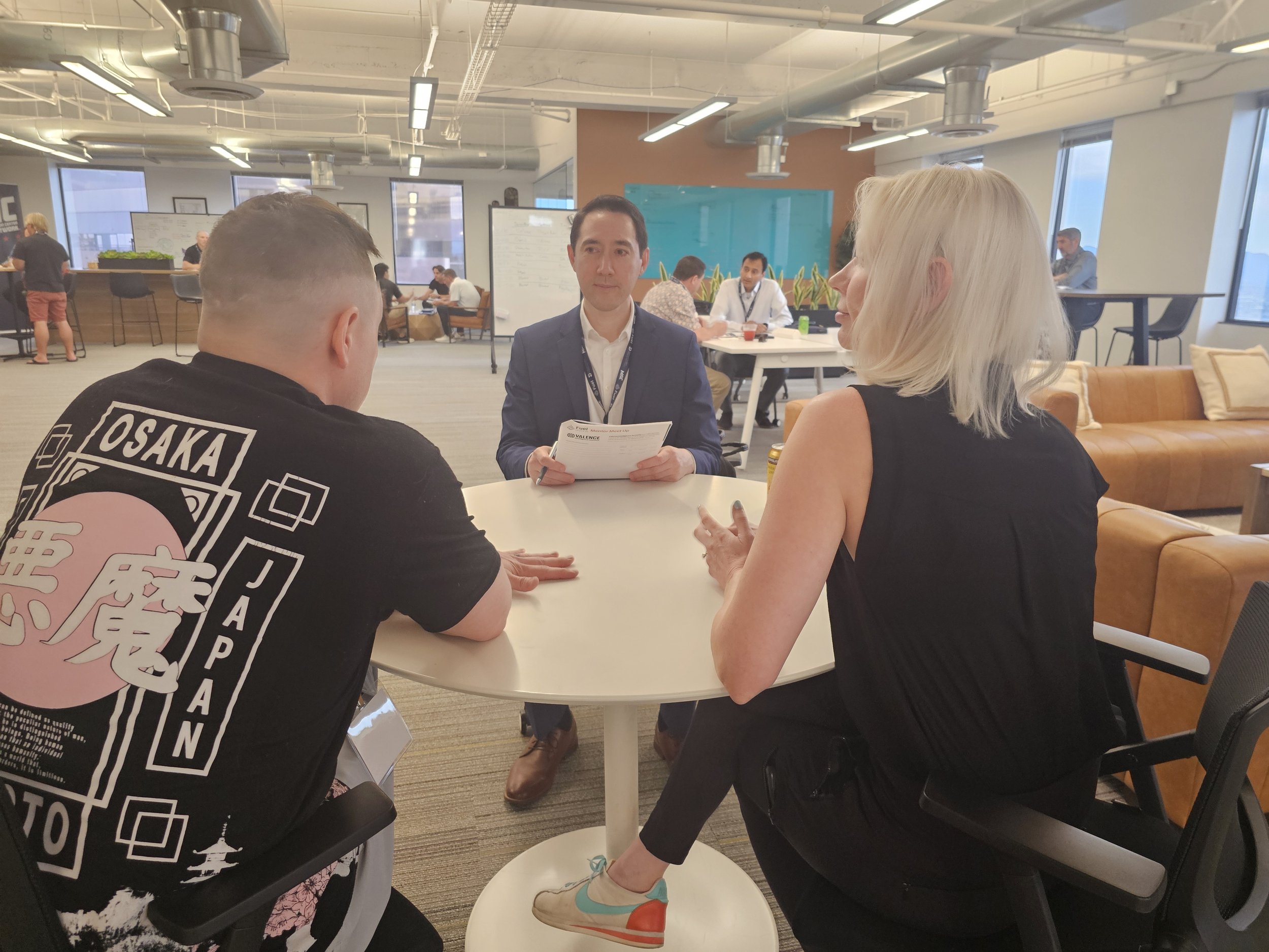 Three people sitting at a round table in an open office, engaged in conversation, with more employees working in the background.