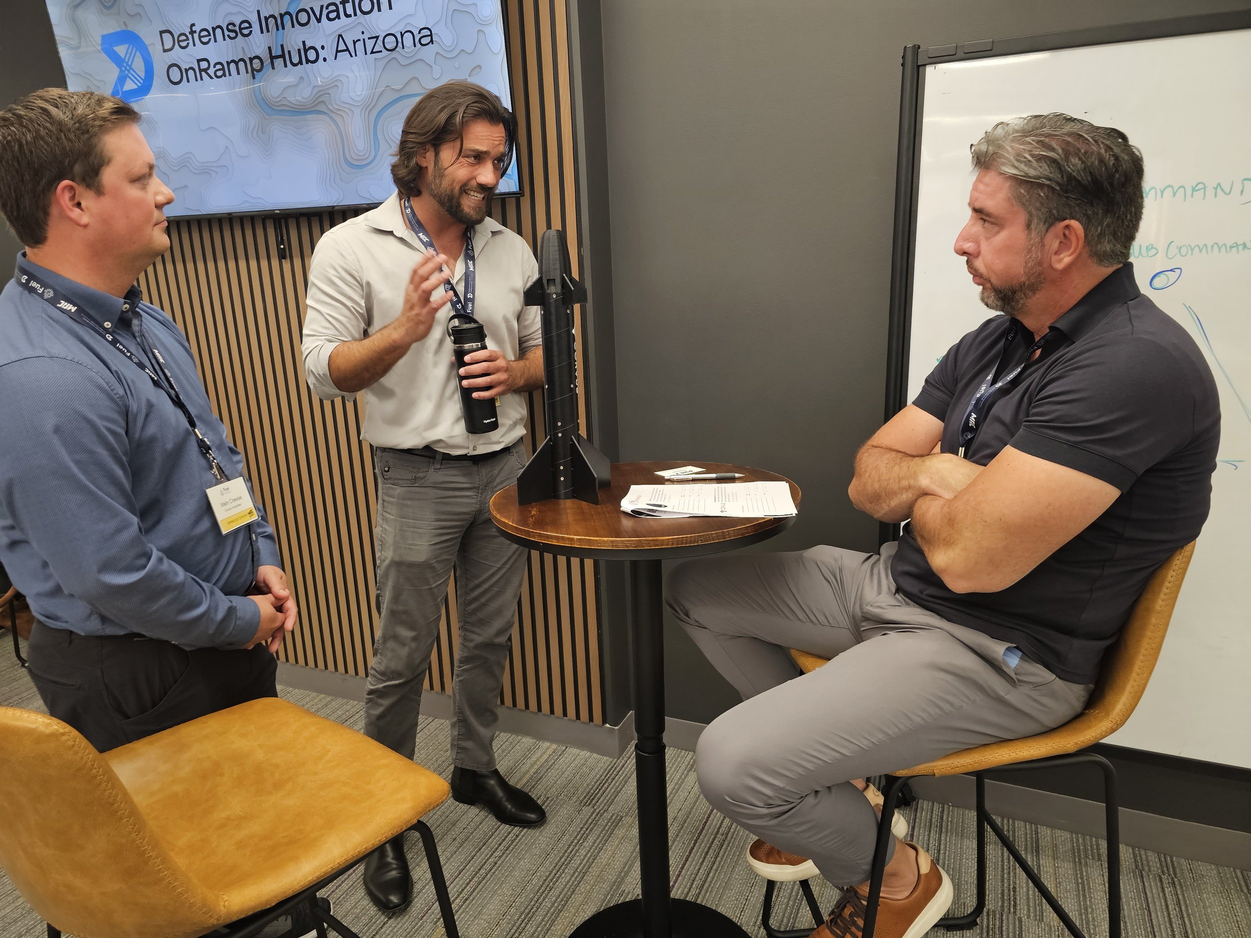 Three men engaged in a discussion in a conference room, one seated with crossed arms and two standing, with a board and a large screen behind them displaying 'Defense Innovation OnRamp Hub: Arizona'.