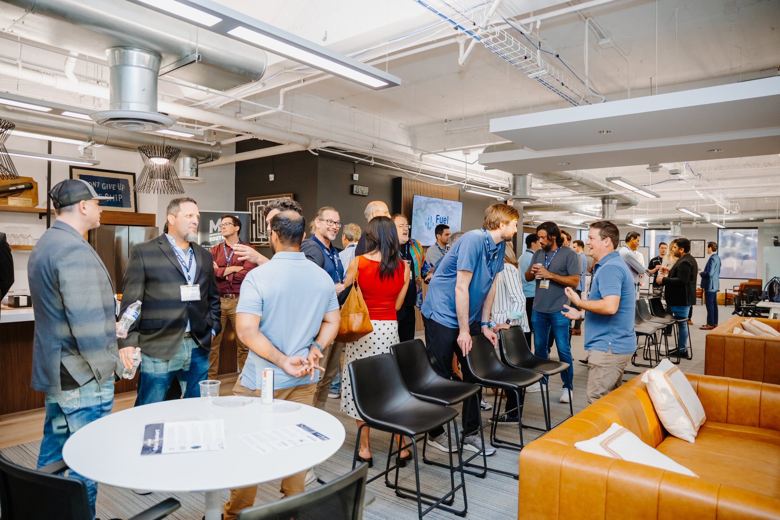 People socializing and chatting in an open-concept corporate or conference space with modern decor, high ceilings, and a large flat-screen TV.