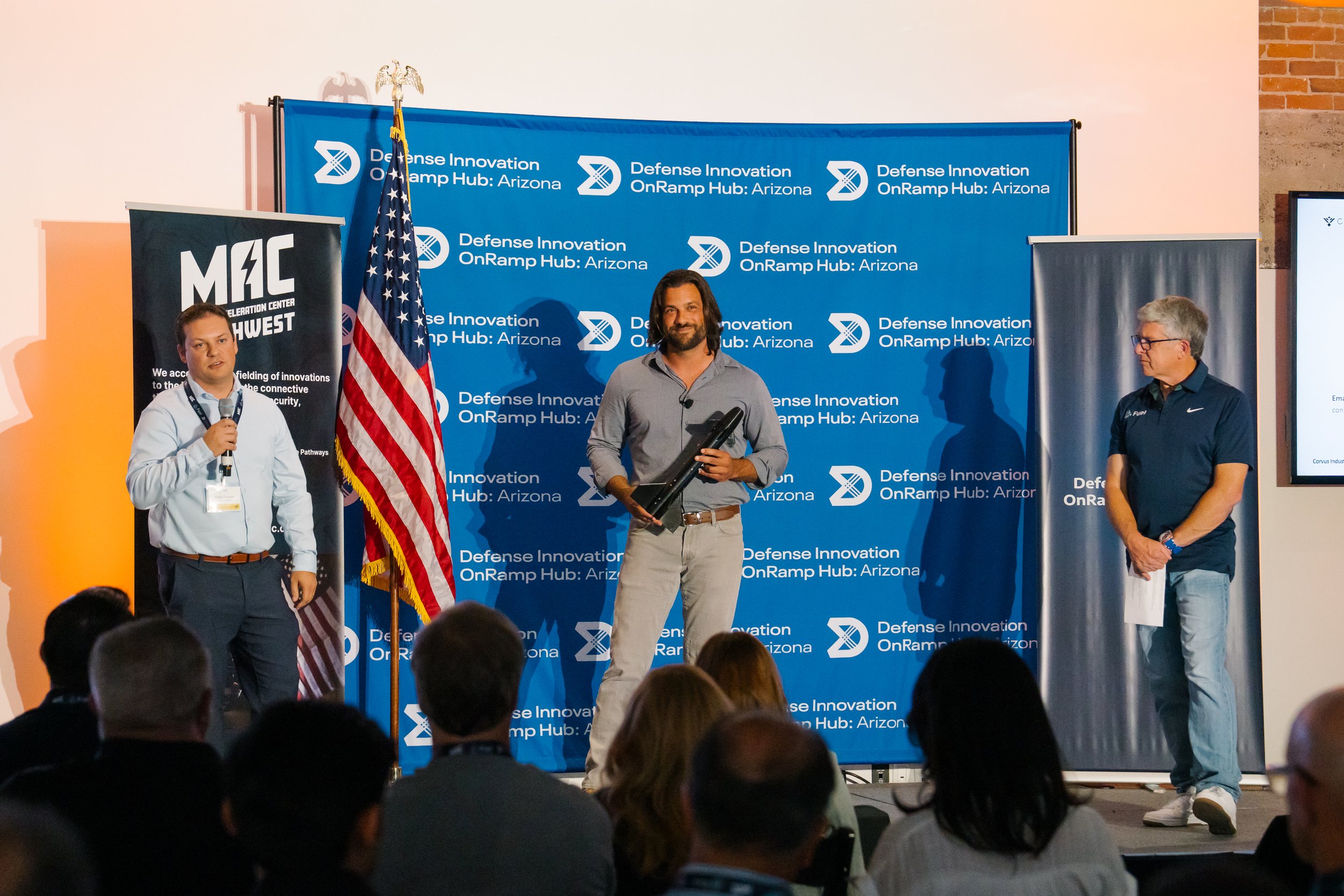 A man holding a black object, possibly a drone or a piece of technology, stands on a stage in front of a blue backdrop with 'Defense Innovation OnRamp Hub: Arizona' written repeatedly. Two other men stand on either side; one with a microphone, and th