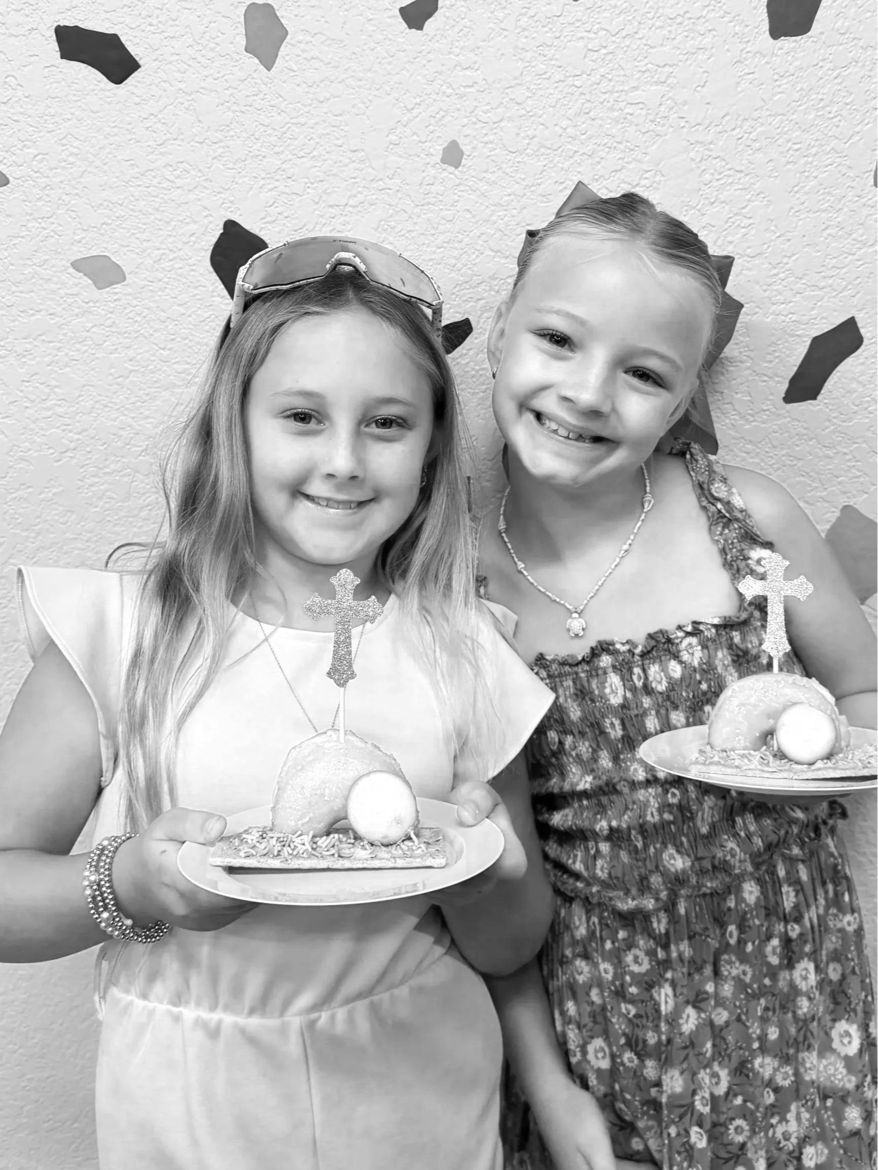 Two young girls smiling and holding plates of cake, with the girl on the left wearing sunglasses on her head and the girl on the right wearing a floral dress and a necklace, in front of a textured wall decorated with confetti and glittery cross ornaments