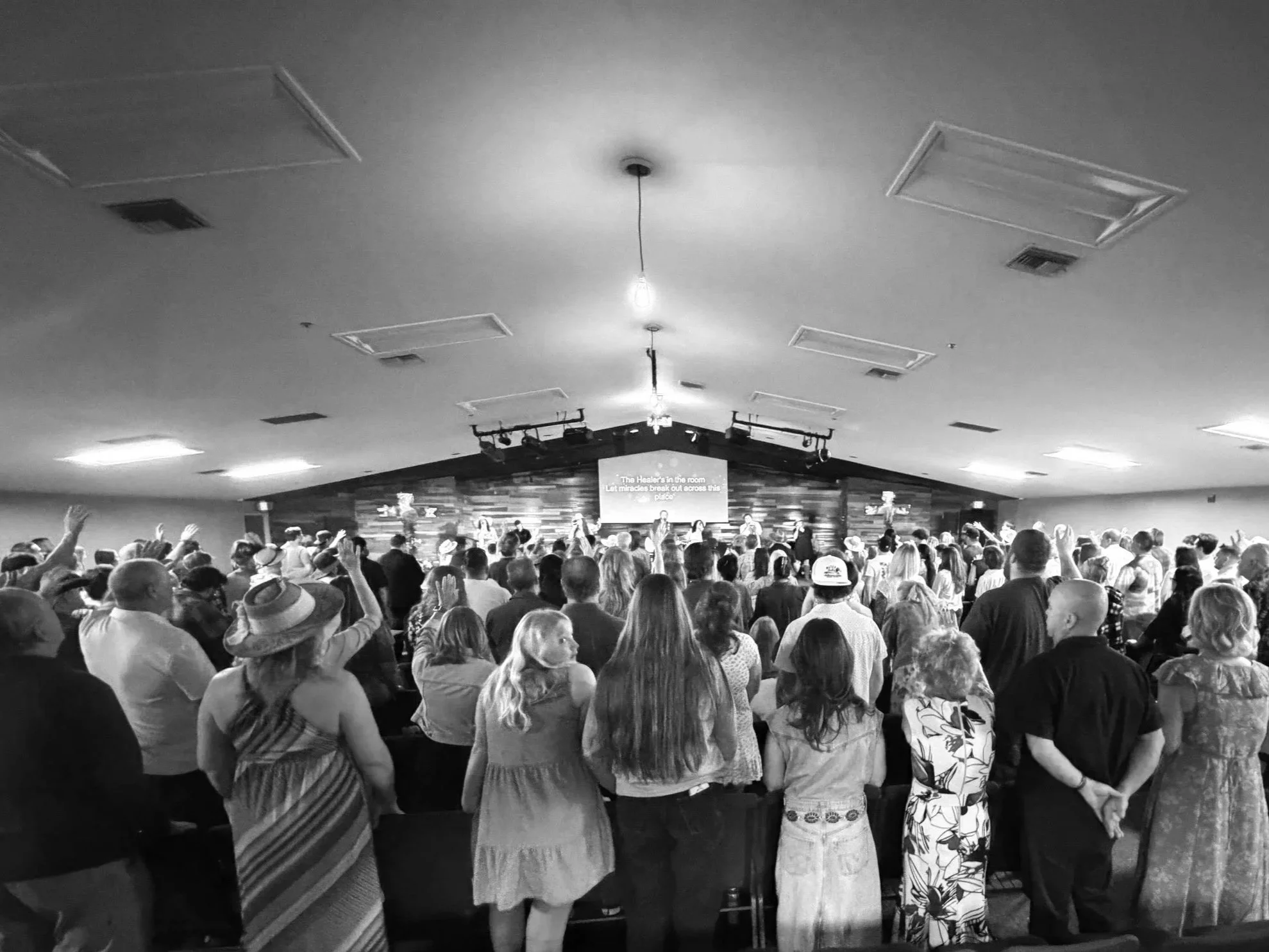 A large group of people gathered in a church or concert hall, facing a stage with a group of performers or speakers. The audience includes men, women, and children of various ages. Some audience members are raising their hands, possibly participating in singing or an activity. The stage has a wooden backdrop, and a sign on the wall reads: 'The Healer's in the room. Let miracles break out across this place.'