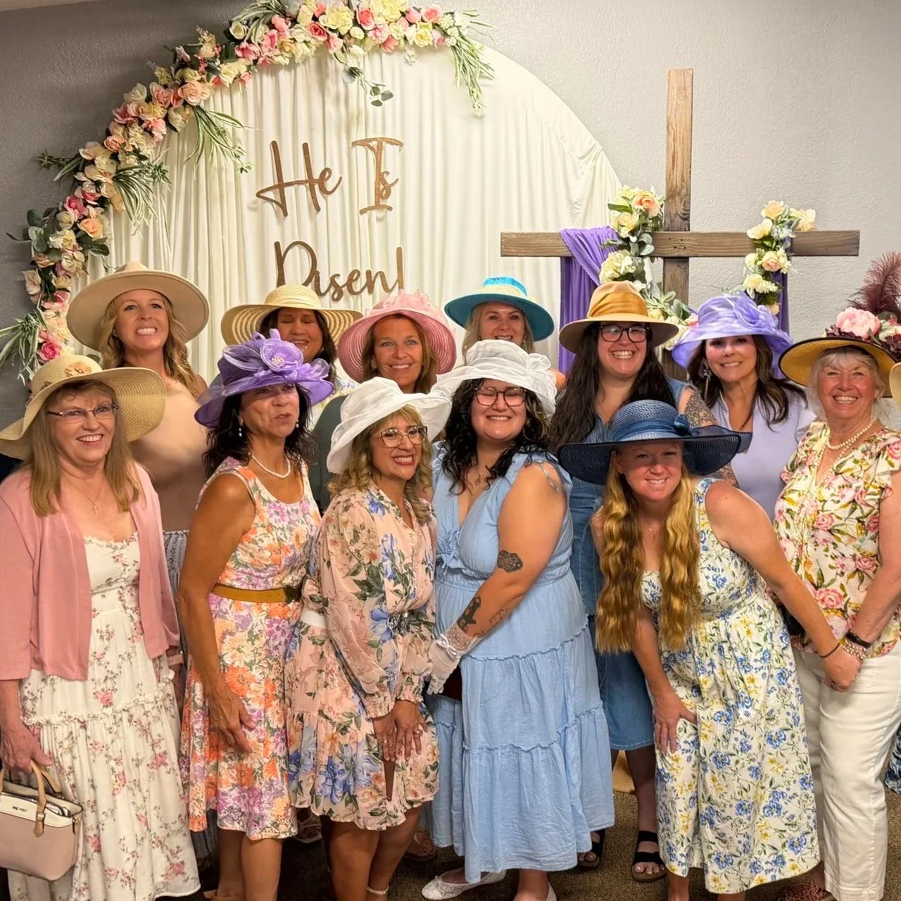 Group of women dressed in floral dresses and large colorful hats, gathered in front of a cross with floral decorations, at a celebration event.