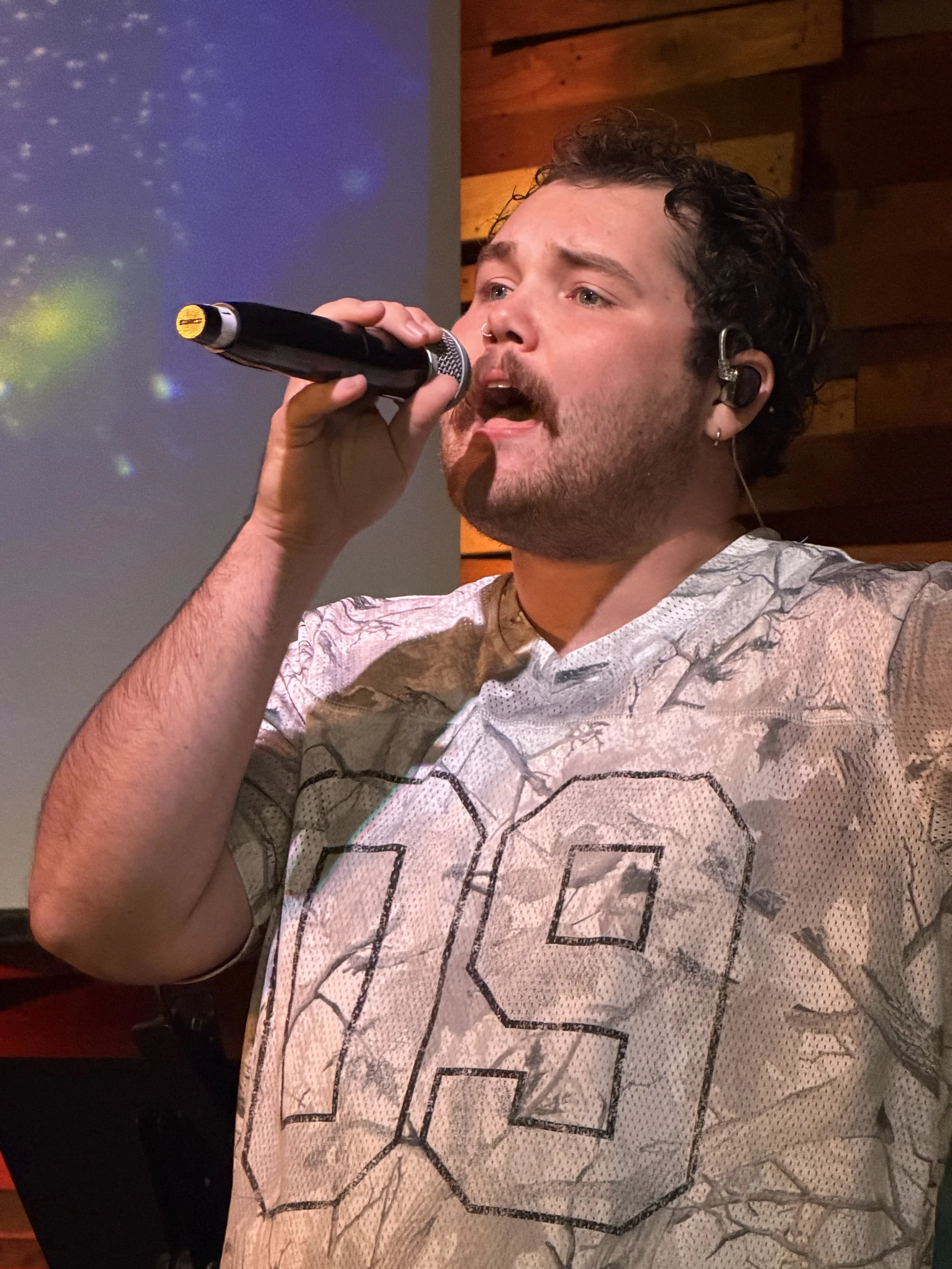 A man with dark, curly hair and facial hair singing into a microphone, wearing an earpiece, t-shirt with a geometric pattern, in church praising god.
