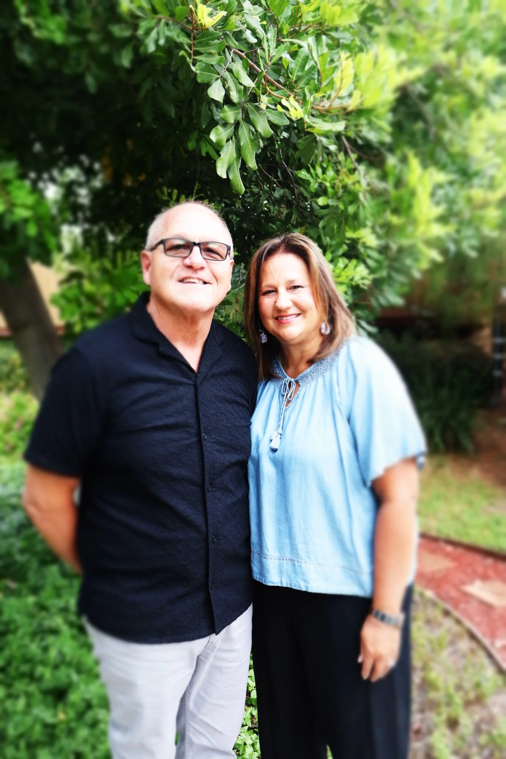 A smiling middle-aged man and woman standing outdoors in front of green trees and bushes, posing for a photo together.