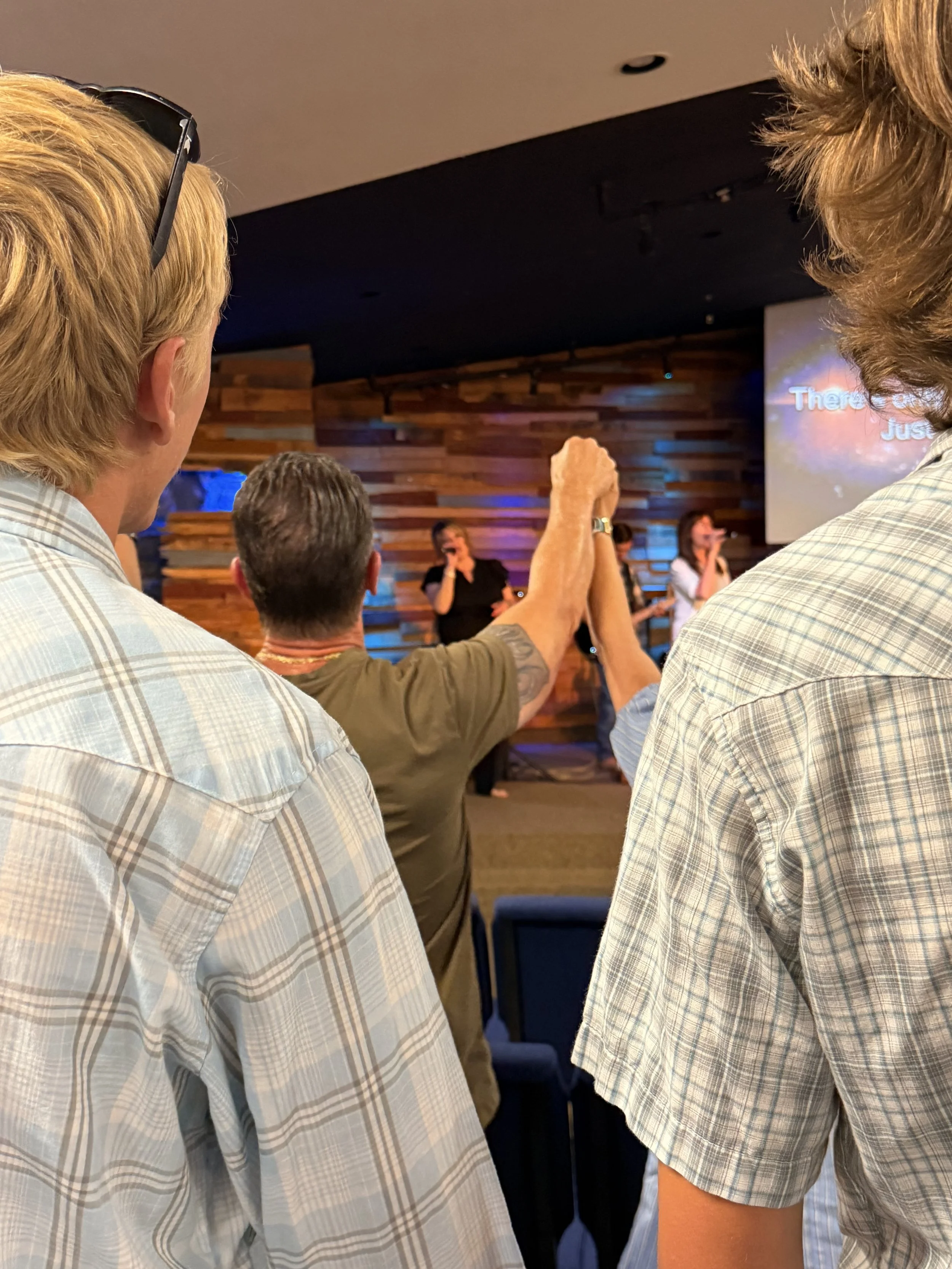 People standing in a room with wooden panel walls, some raising their hands, others holding microphones, with a large screen displaying words in the background.