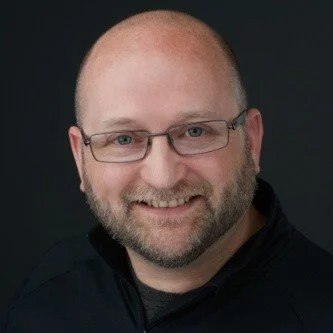 A headshot of the founder - A white male with glasses, bald, with a beard.
