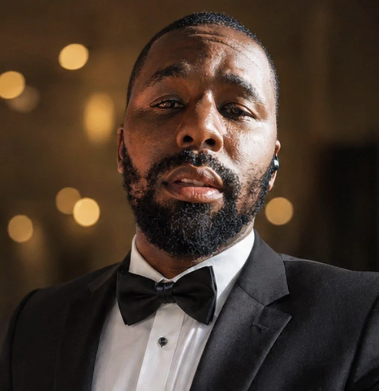 A close-up of a man dressed in a black tuxedo with a bow tie, with a serious expression, short hair, and a beard, standing in a dimly lit setting with blurred circular lights in the background.