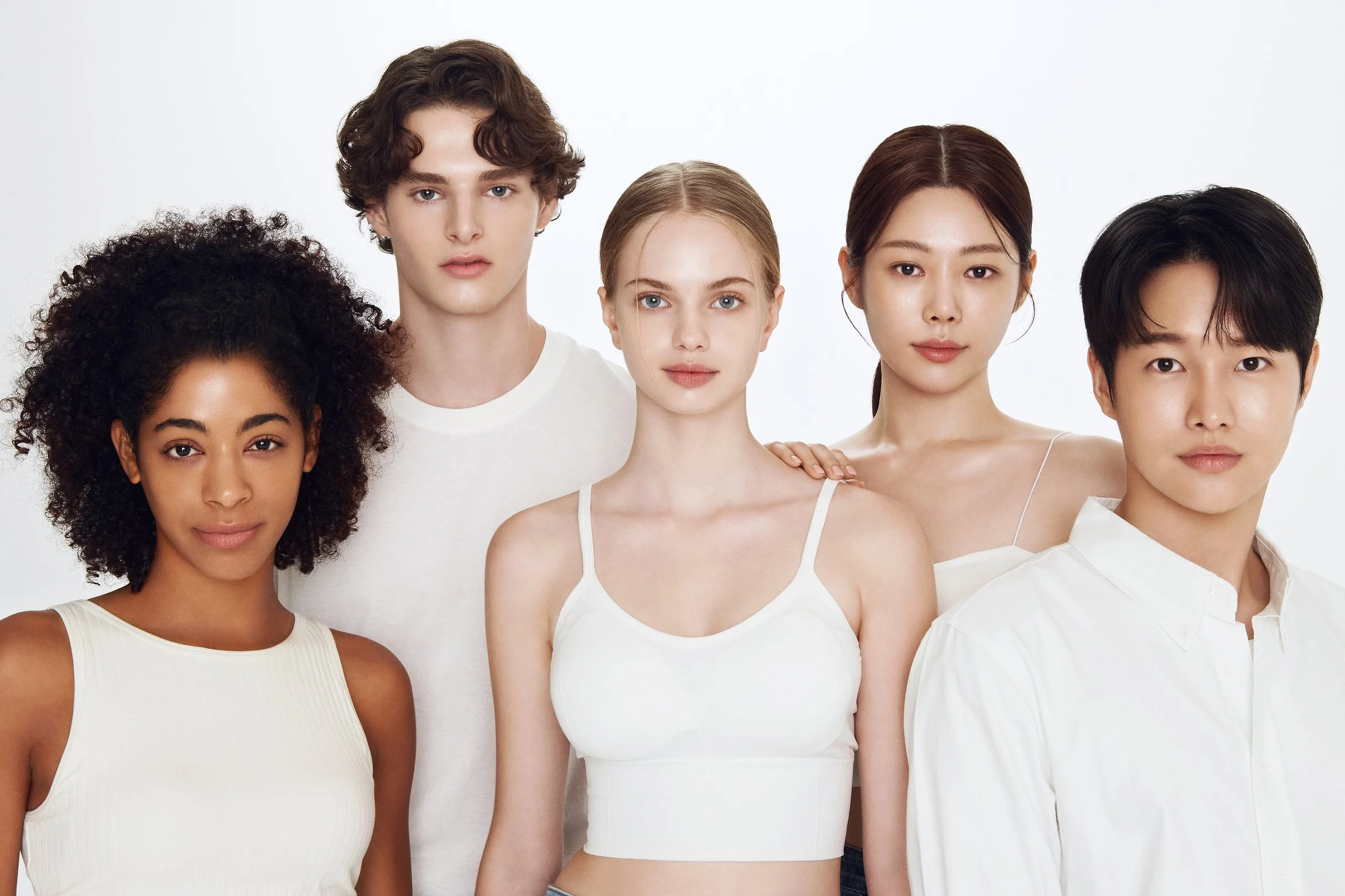 Groupe de cinq jeunes adultes diversifiés, posant ensemble contre un fond blanc, tous portant des vêtements blancs ou neutres.