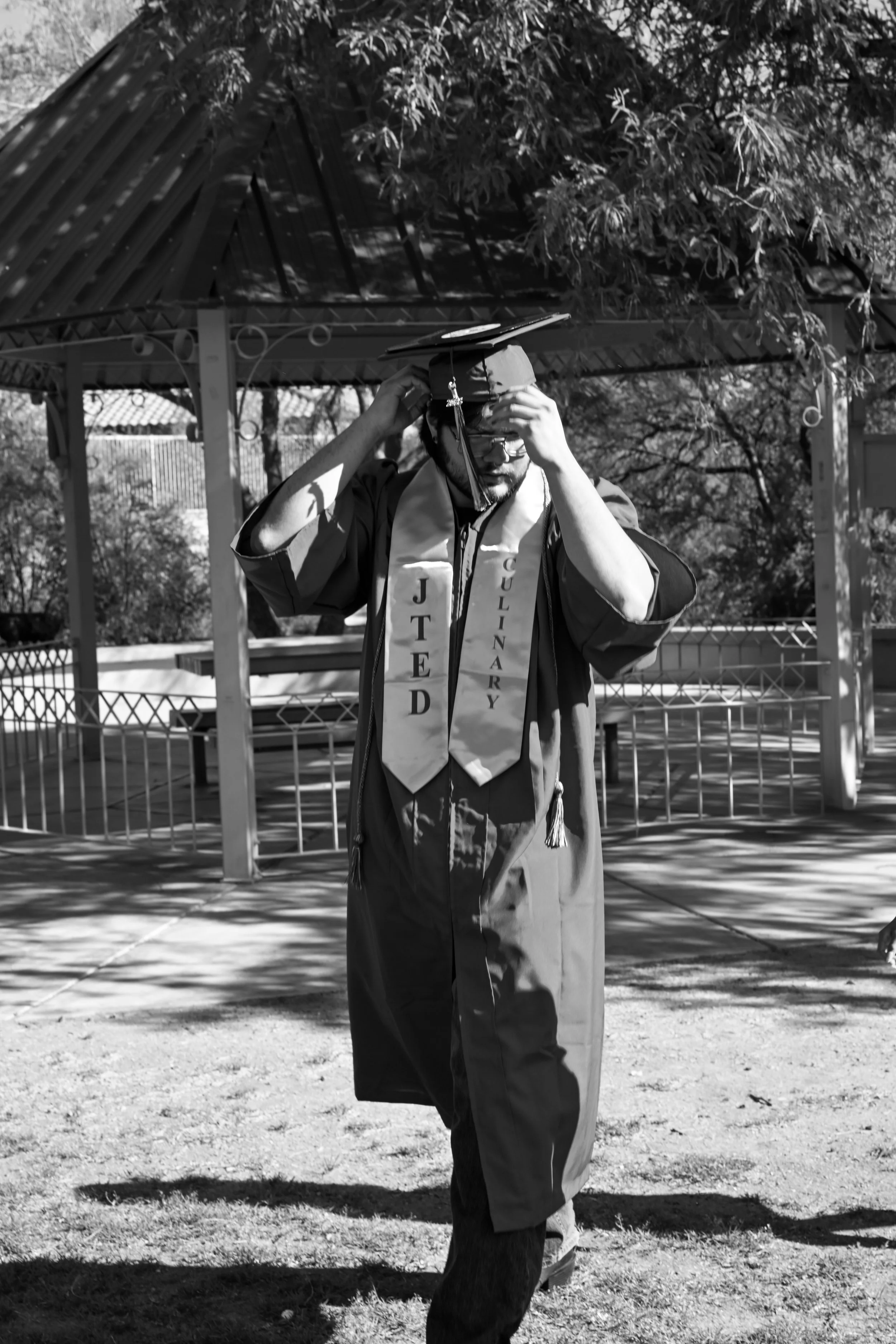 A man in graduation gown and cap adjusting his cap, standing outdoors near a gazebo and trees, with a graduation stole reading "JTED Culinary".