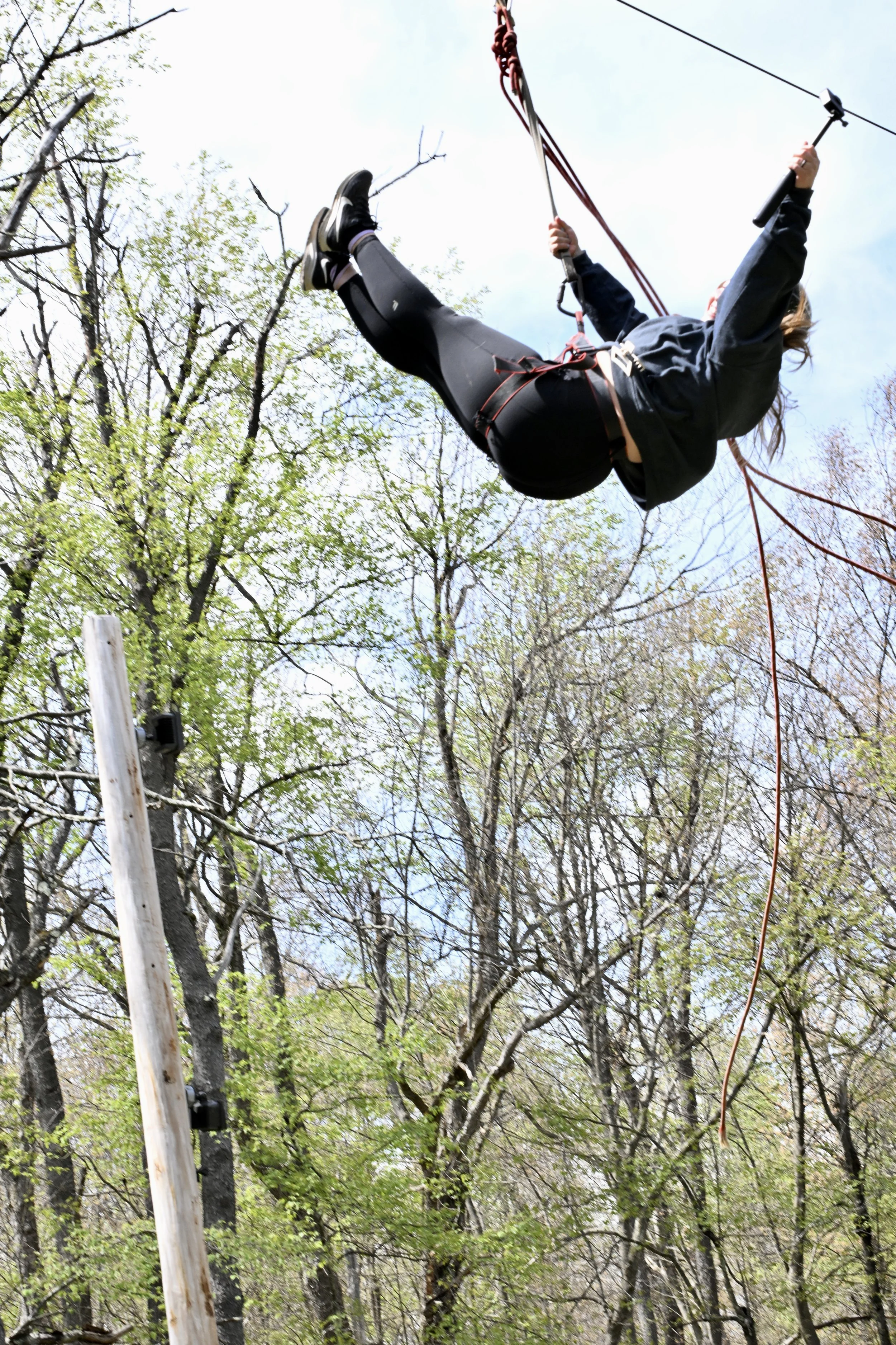 Person wearing black clothing and a harness swinging high above the ground in a tree-filled outdoor setting.