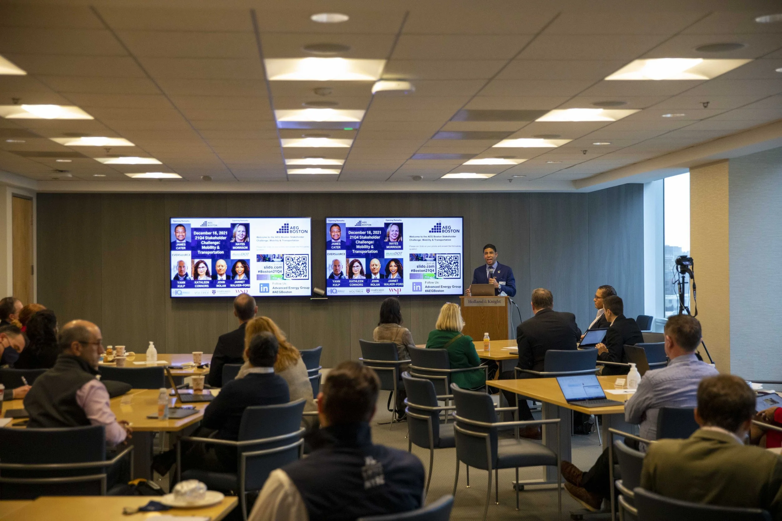 A speaker presenting at a conference in a conference room with an audience seated at tables, large screens displaying information behind him, and a camera set up to record the event.