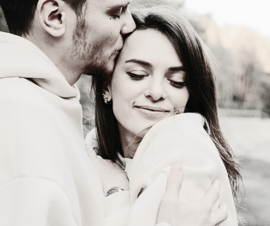 A black and white close-up photo of a man gently kissing a woman on the forehead while embracing her, both wearing hoodies and smiling softly.