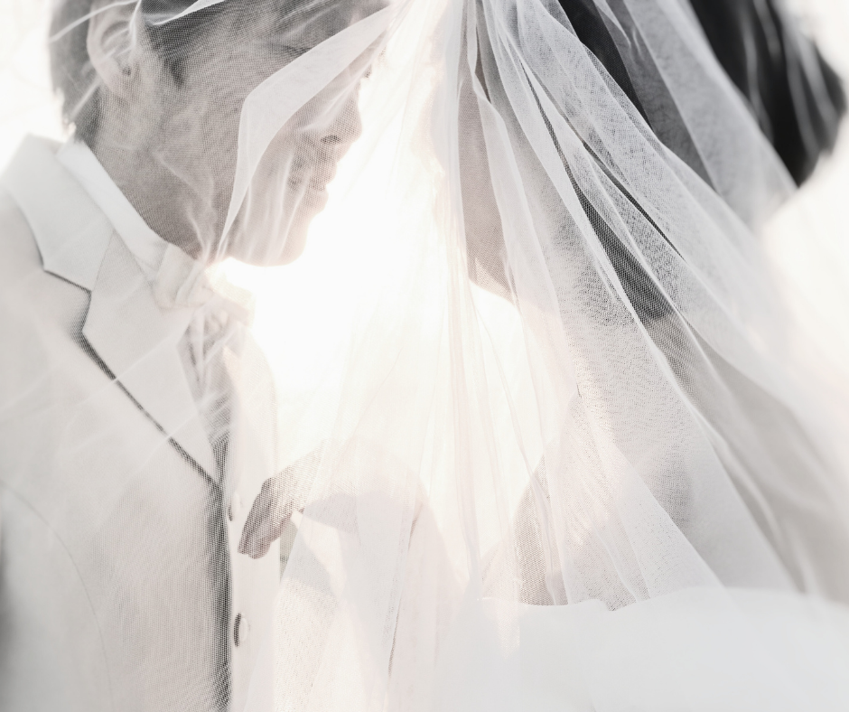 A bride and groom standing close together, draped in a sheer veil, with soft backlighting creating a romantic and ethereal atmosphere.