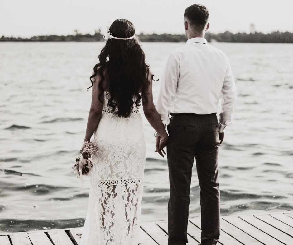 A couple dressed in wedding attire standing hand in hand on a dock by the water, facing away from the camera, with a calm body of water and distant shoreline in the background.