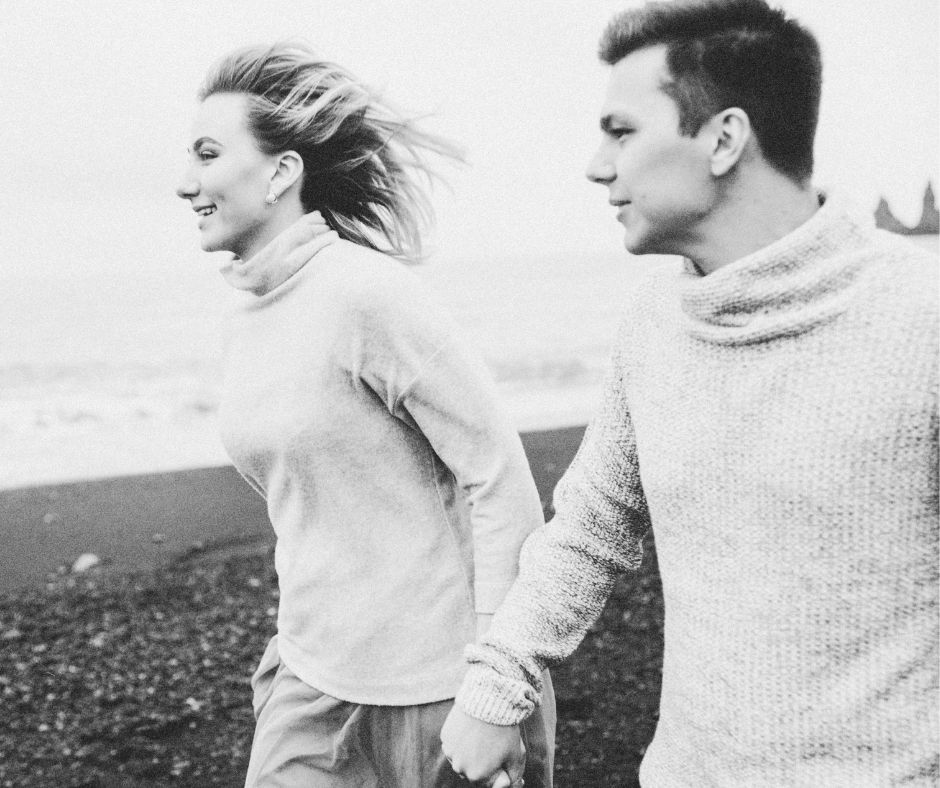 A black and white photo of a smiling woman holding hands with a man on a beach, both facing to the right, with the woman's hair blowing in the wind.