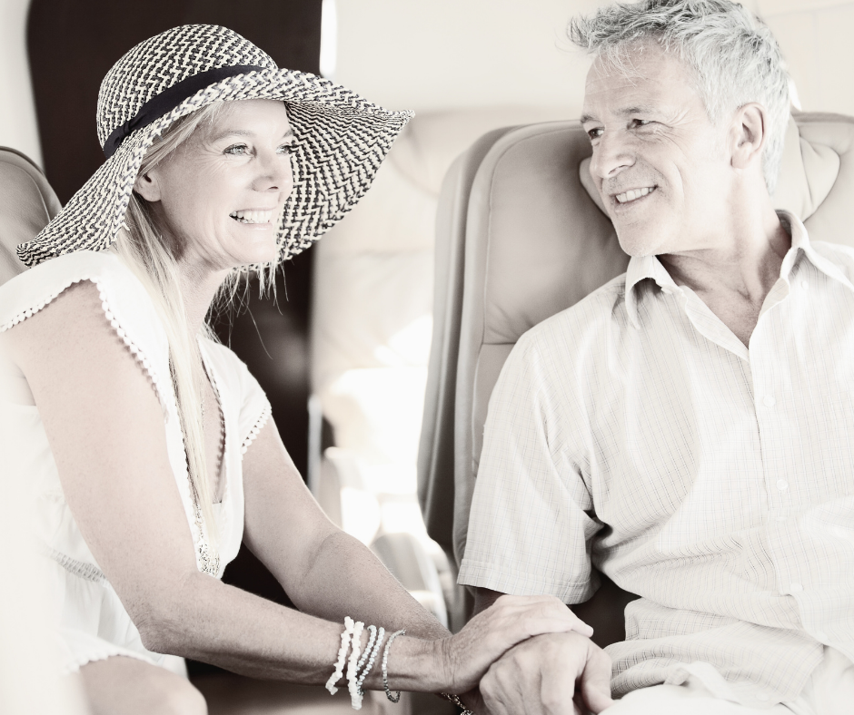 An elderly woman and man sitting in airplane seats smiling and holding hands, engaged in a joyful conversation.