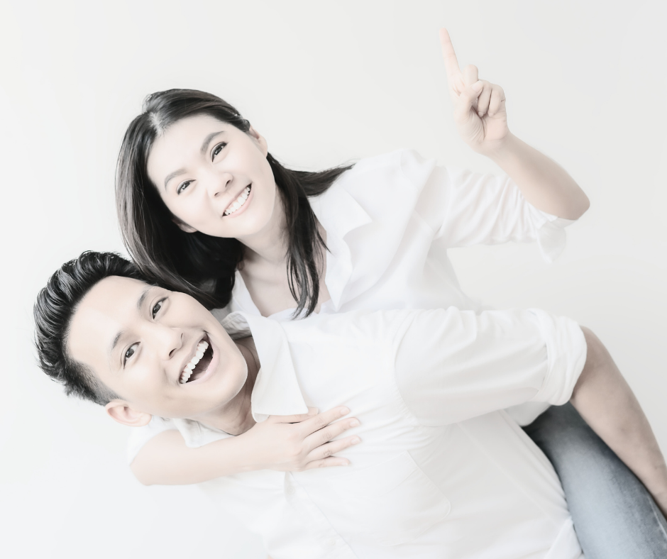 A smiling woman giving a piggyback ride to a man, both dressed in white shirts, against a white background.