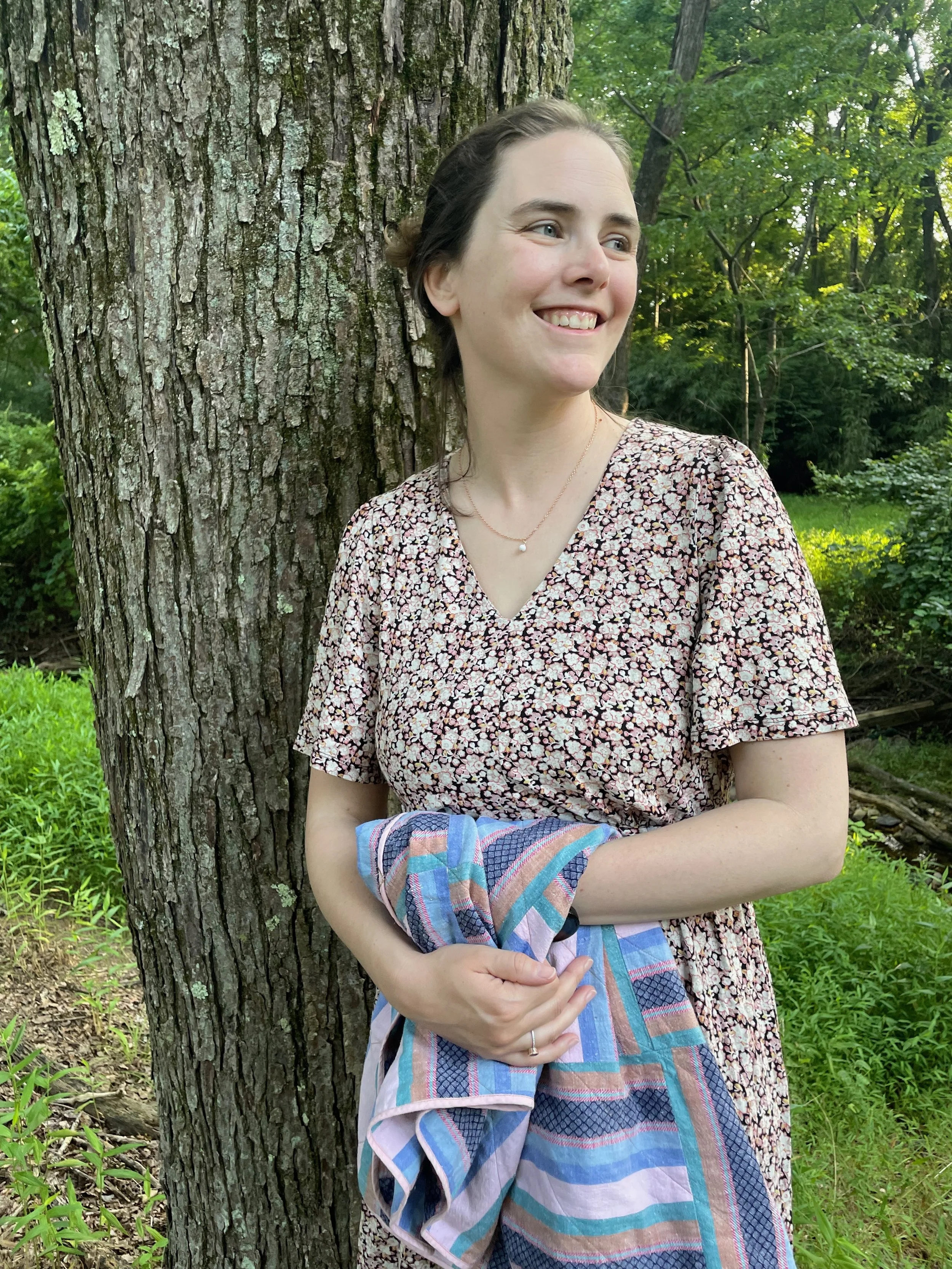 A woman standing next to a tree in a green outdoor setting, smiling and holding a plaid blanket.