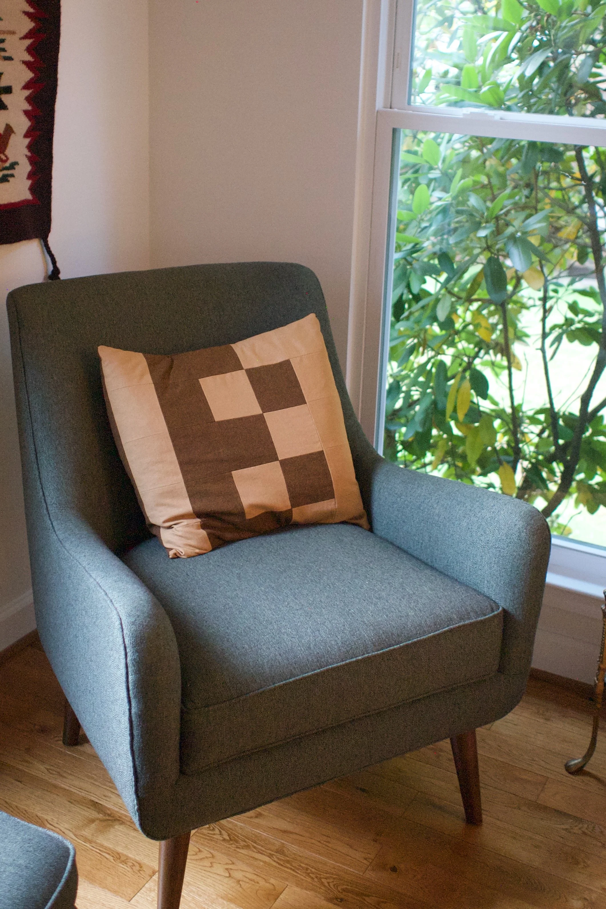 A brown and beige patterned throw pillow rests on a chair. A dark green mid-century modern armchair with wooden legs, positioned on a wooden floor, next to a window with green leafy plants outside.