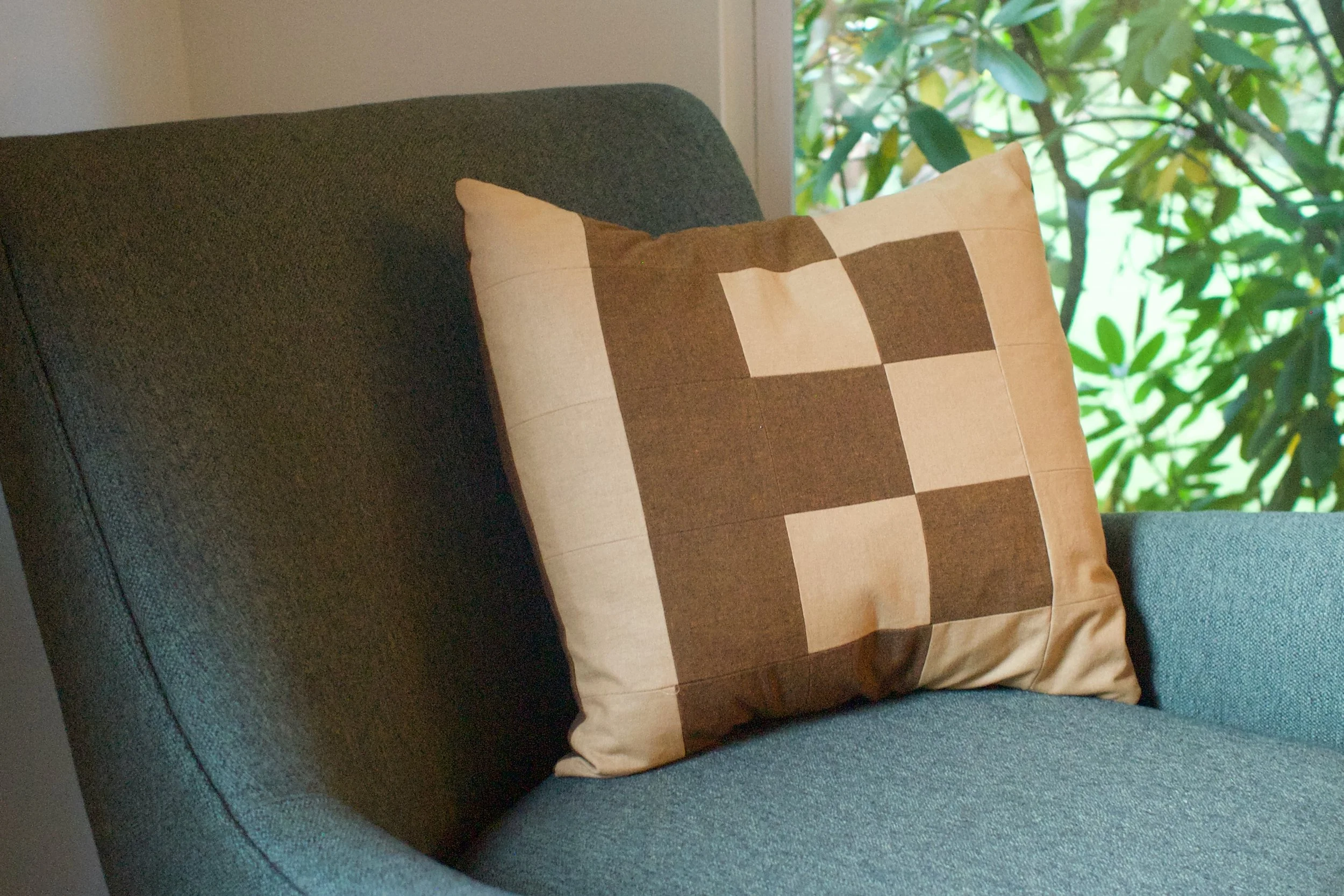 A decorative pillow with a geometric pattern in shades of beige and brown resting on a dark green armchair near a window with green foliage outside.