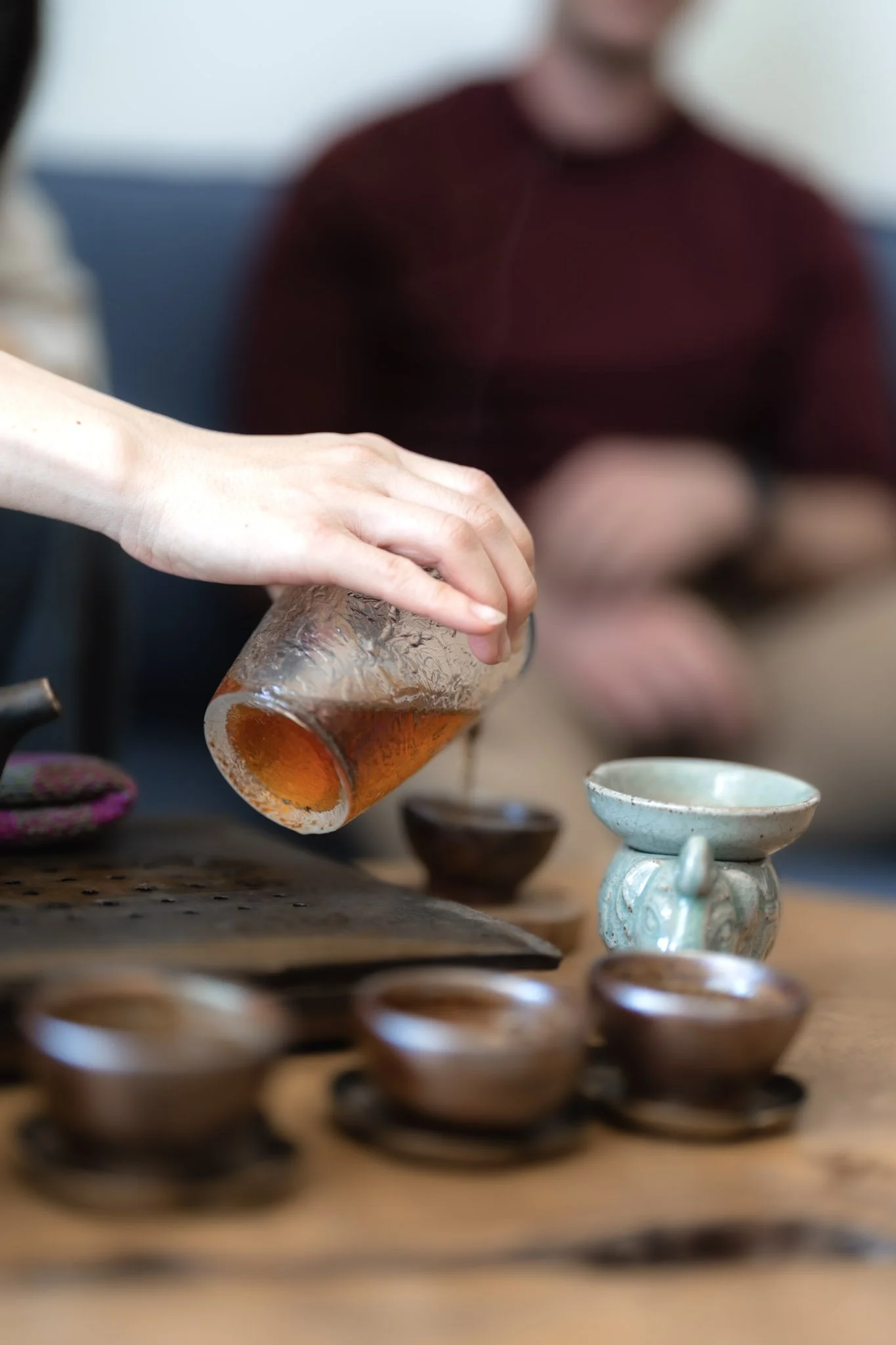 Person pouring tea into a small ceramic cup on a wooden table, with other cups and tea utensils nearby, people in the background.