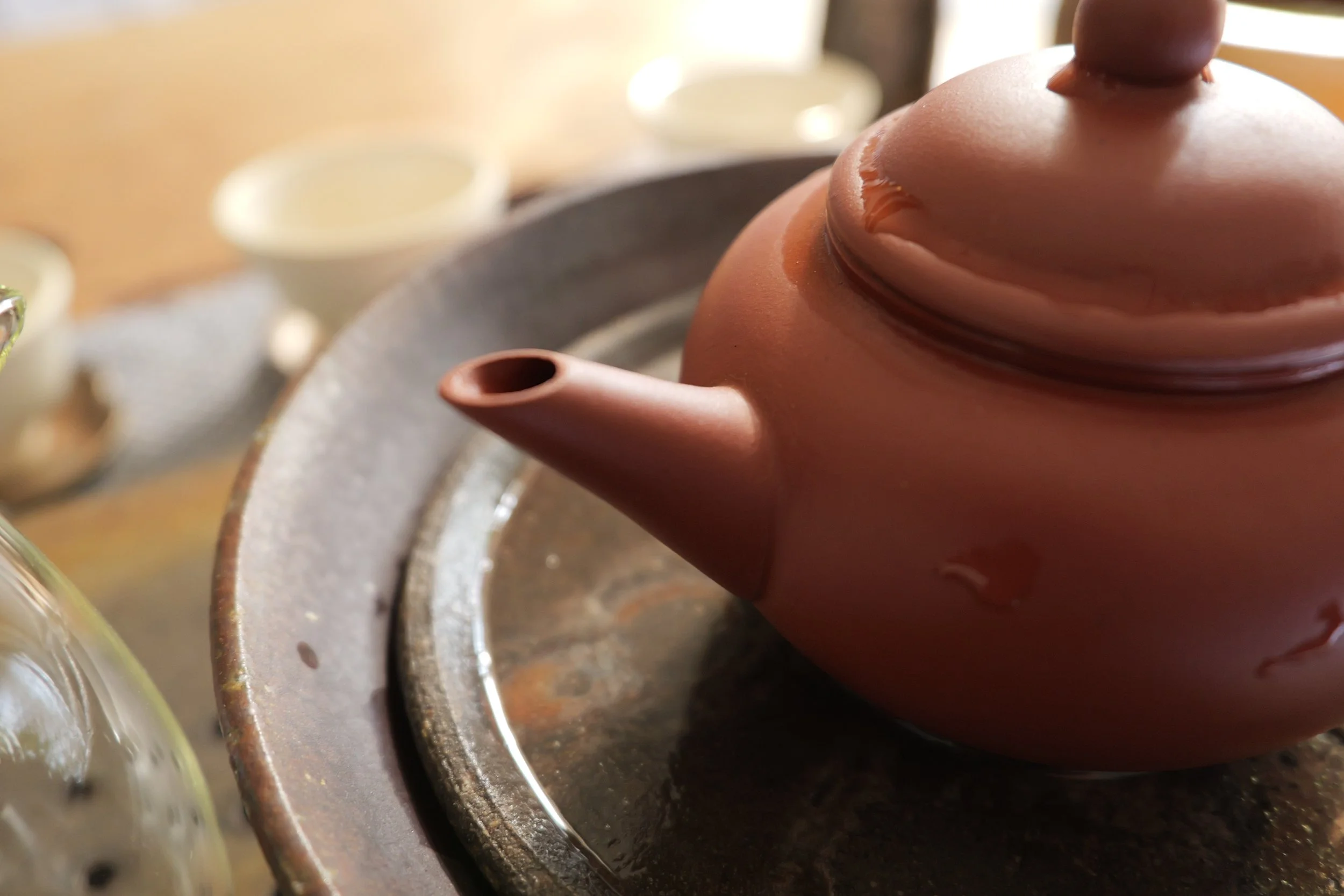 Close-up of a brown clay teapot on a metallic tray, with blurred tea cups and teapot in the background.