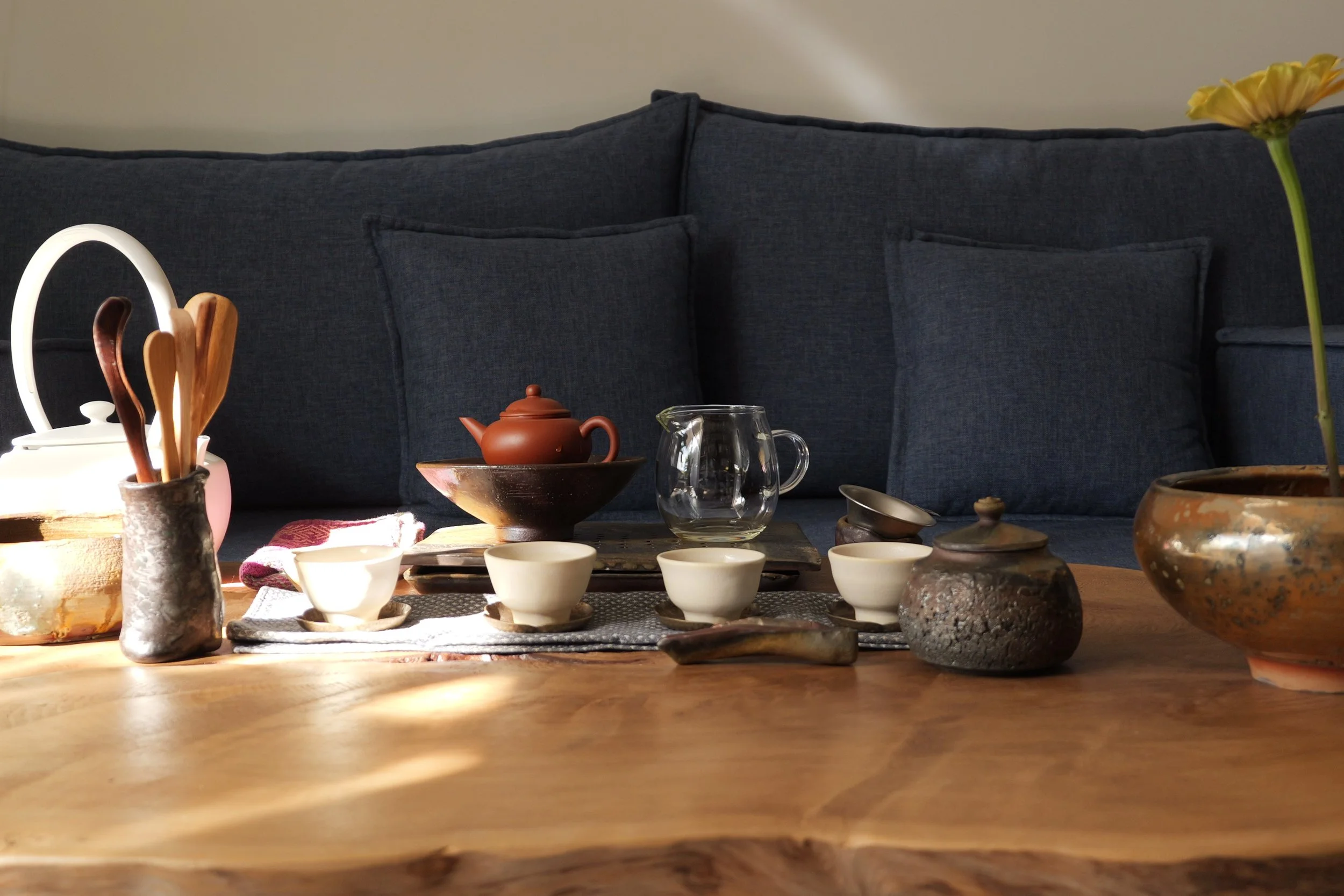 Tea set with teapot, cups, and utensils on a wooden table in front of a dark blue couch with cushions.