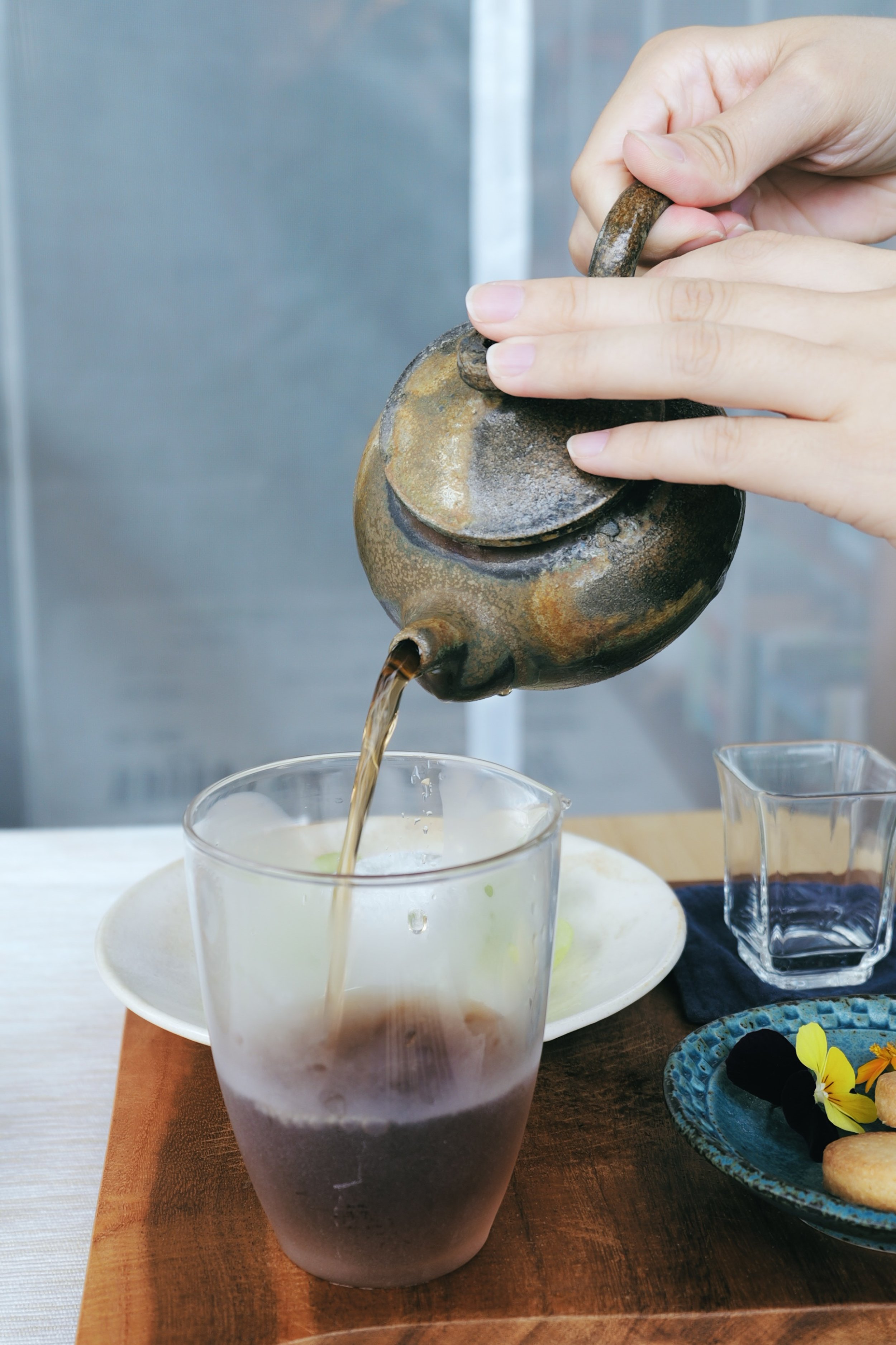 Person pouring tea from a teapot into a glass cup on a wooden table.