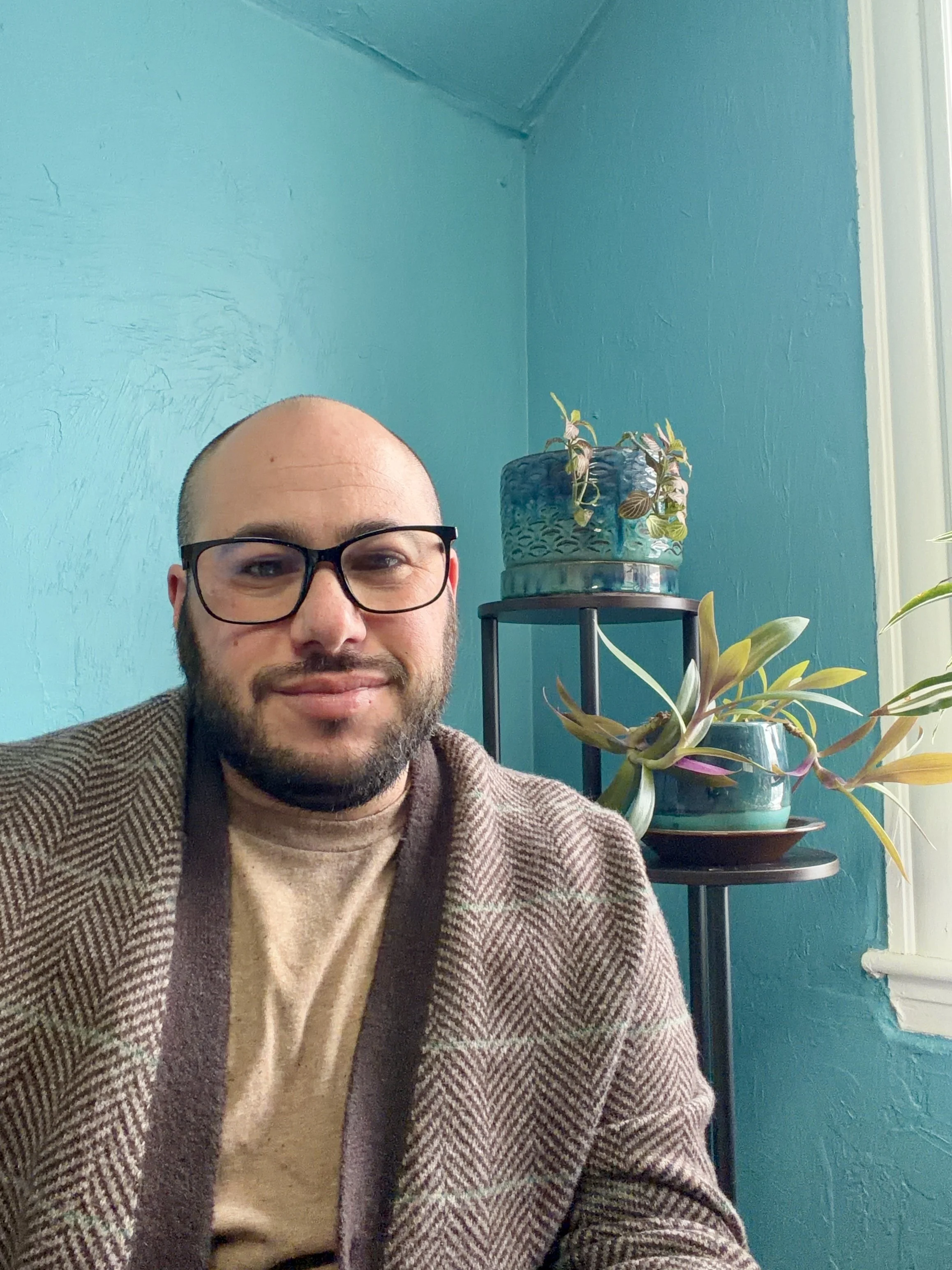 A smiling man with dark hair, a beard, white skin, glasses, and earrings wearing a striped brown and tan cardigan against a blue-colored wall with some plants on a stand in the background.