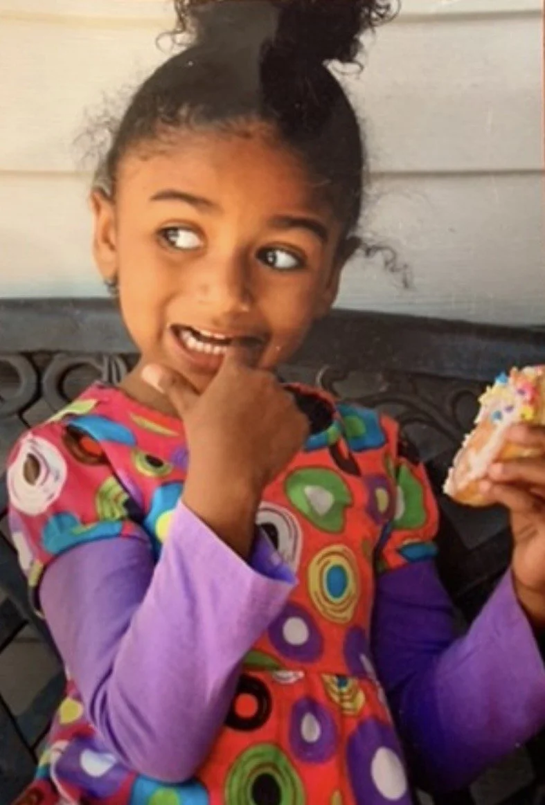A photo of a multi-ethnic little girl wearing a colorful polkadot dress with purple sleeves while holding a donut with sprinkles.