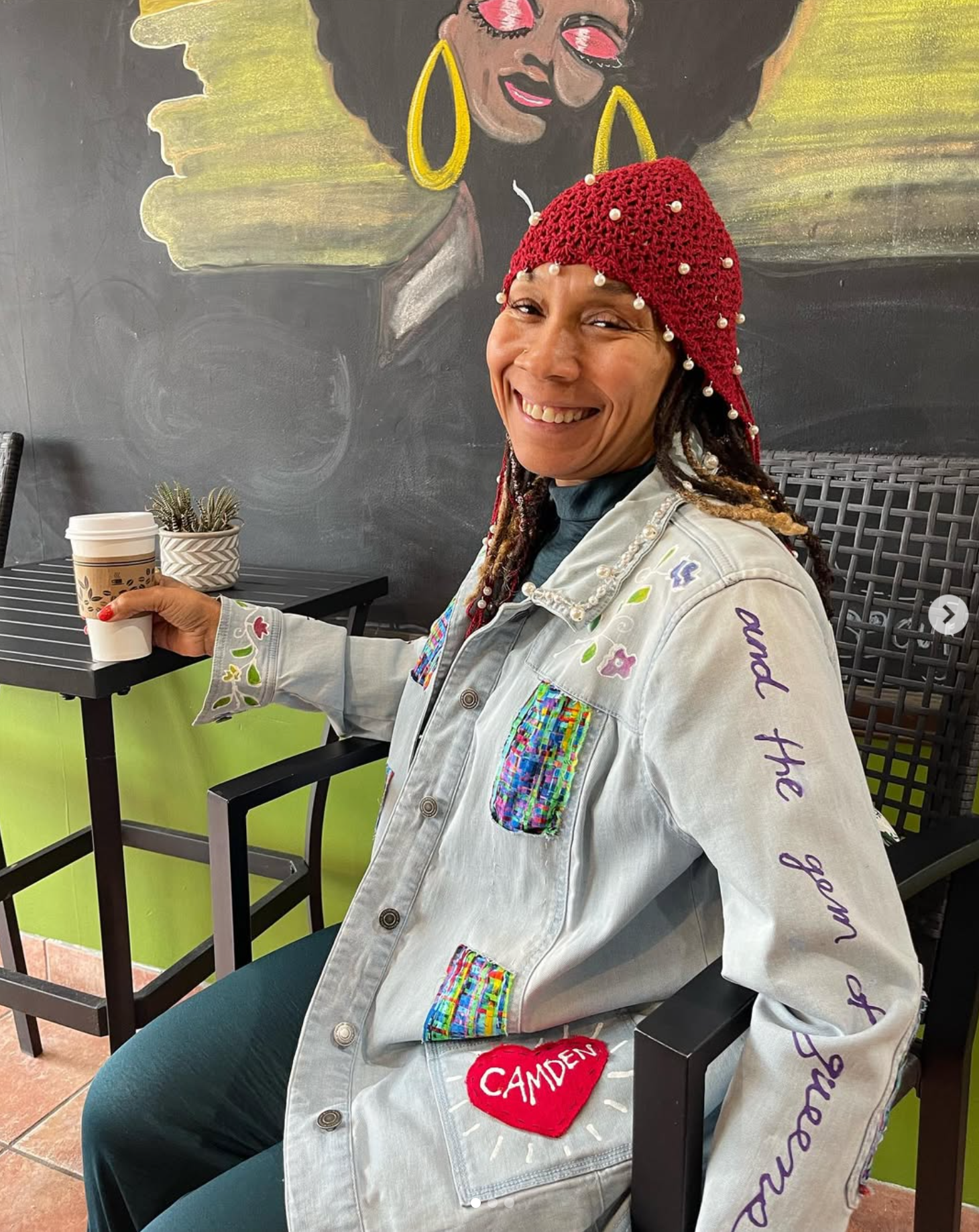 A Black woman wearing a red beanie head covering and a hand painted jacket adorned with pearls, and other hand-sewn patches sitting in a coffee shop
