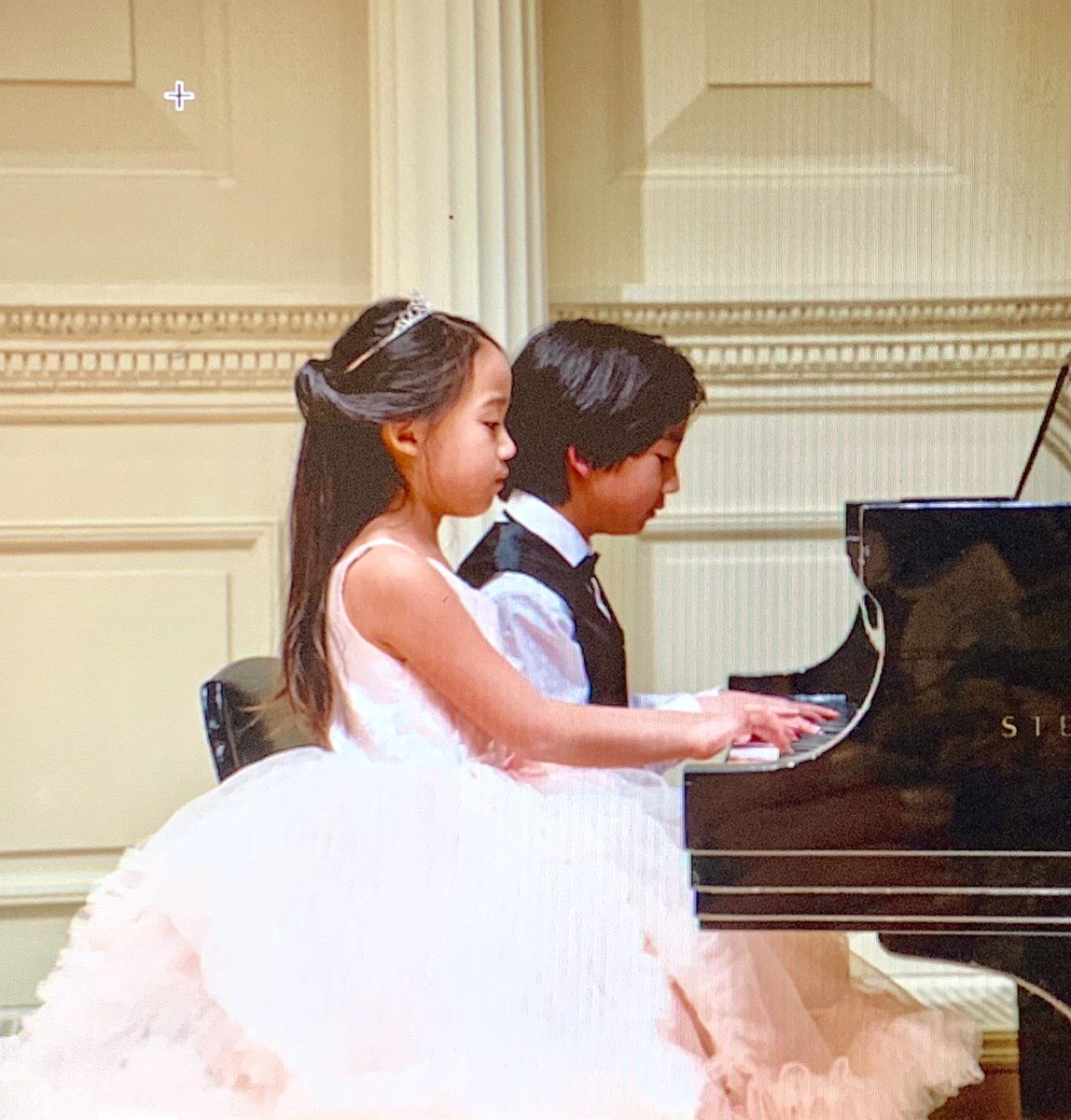 A photo of an Asian girl wearing a white, princess style, fluffy dress and tiara sitting beside her brother playing piano.