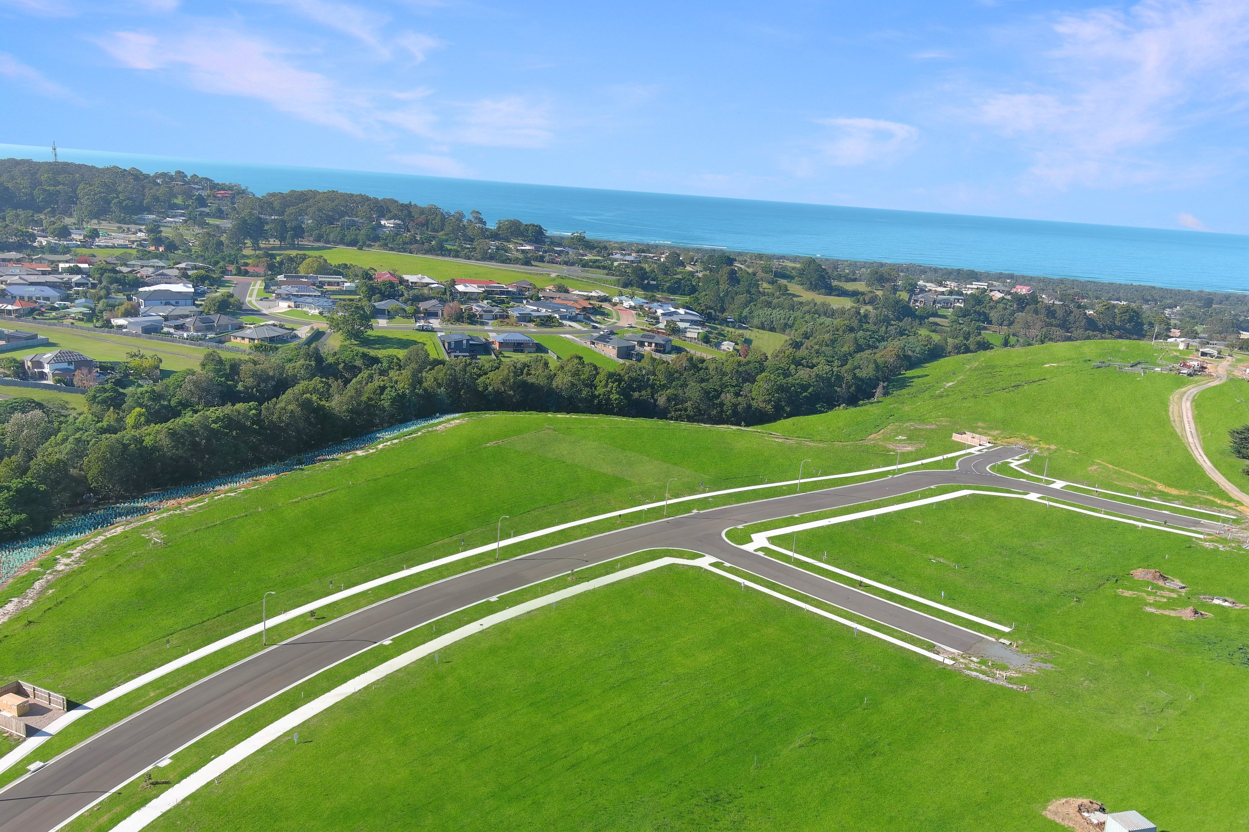 Drone aerial view of Coastal View Estate Stage 3 in Lakes Entrance VIC, showing newly built roads, kerb and footpaths with green rolling hills and ocean views in the background, highlighting titled land for sale and coastal living near the beach.