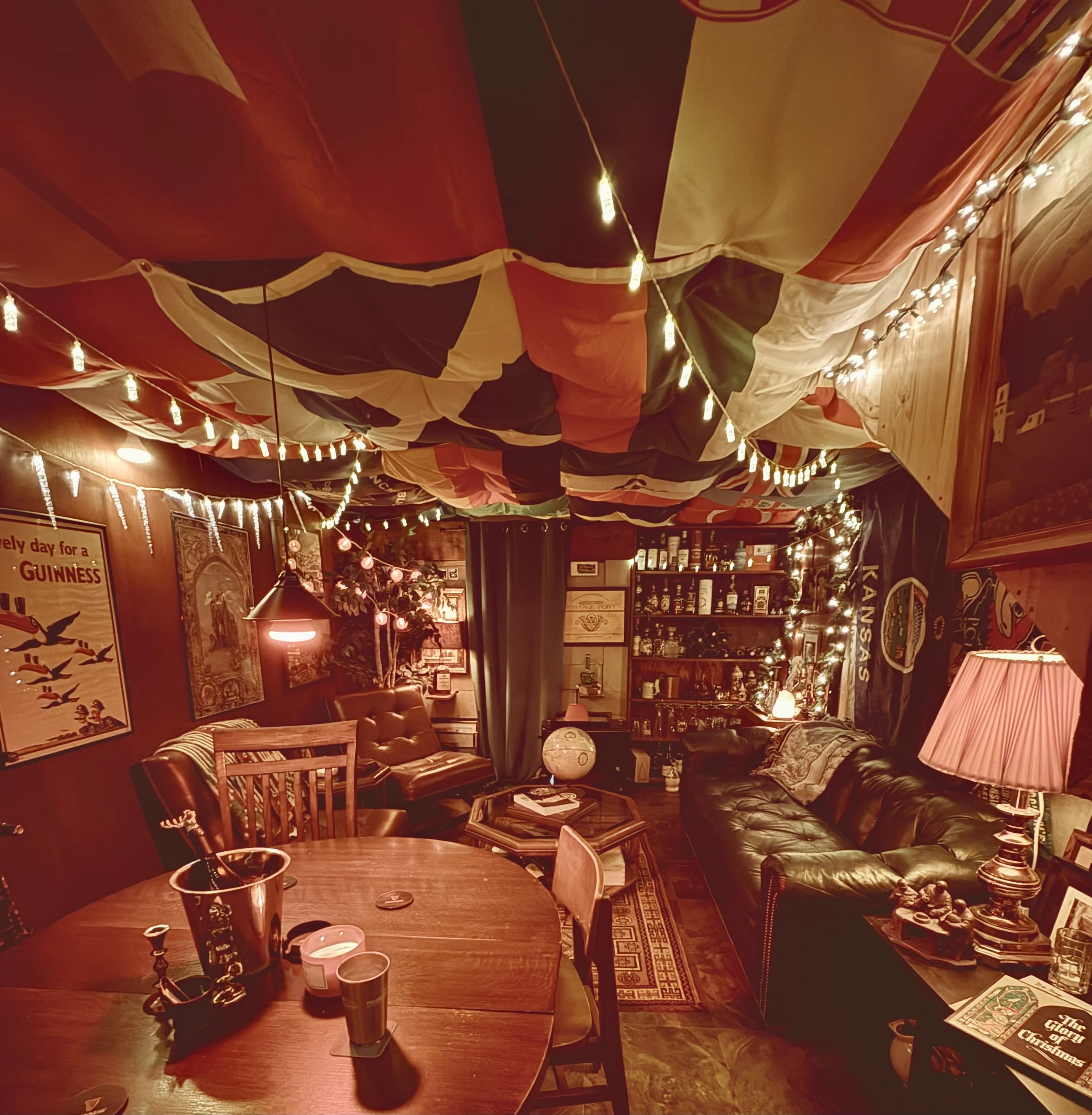 Cozy living room decorated with string lights, batik fabric ceiling, vintage posters, a round wooden table with candles, a leather sofa, a bar shelf with bottles, and warm lighting.