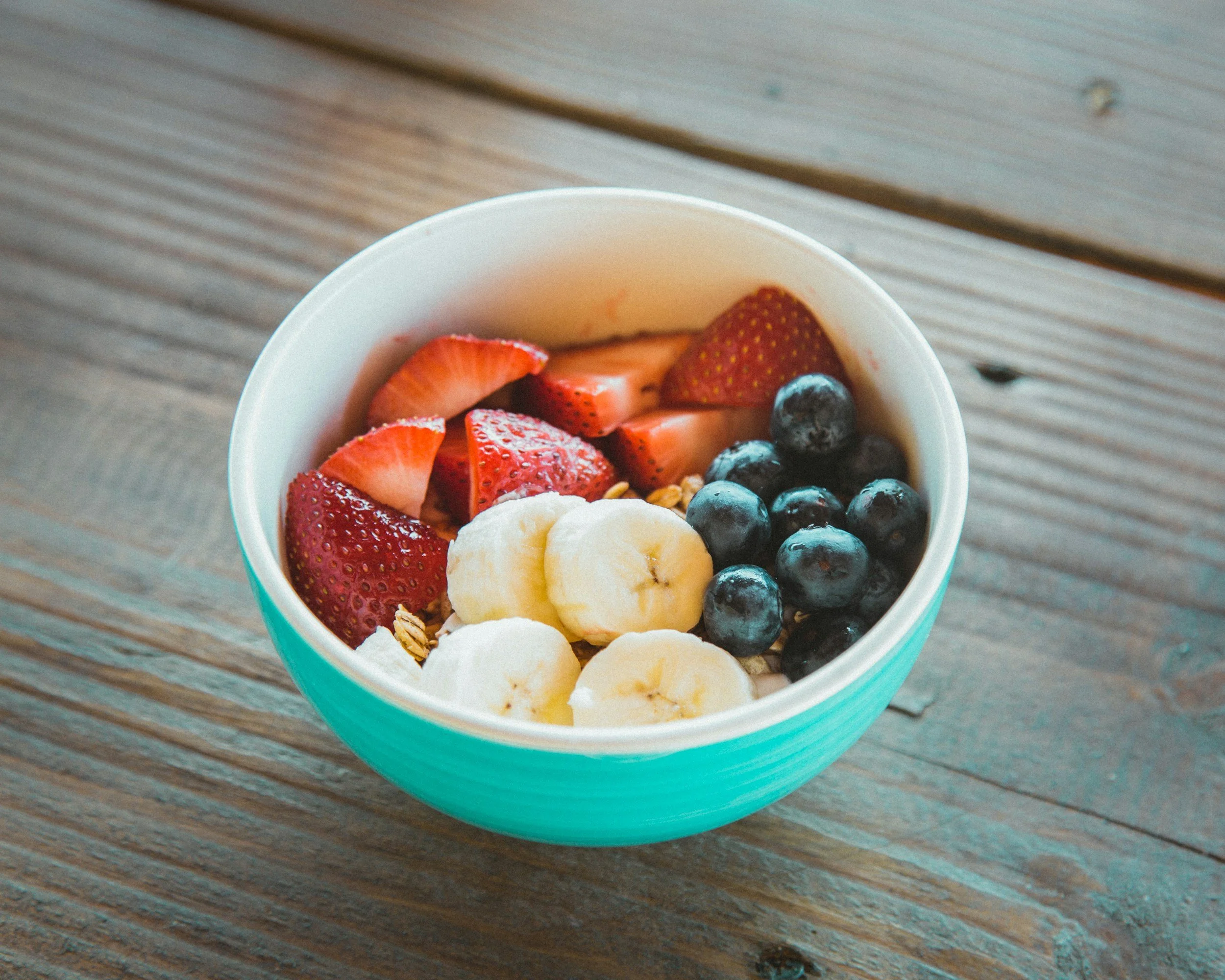 A bowl of yogurt topped with sliced strawberries, blueberries, banana slices, and granola on a wooden surface.