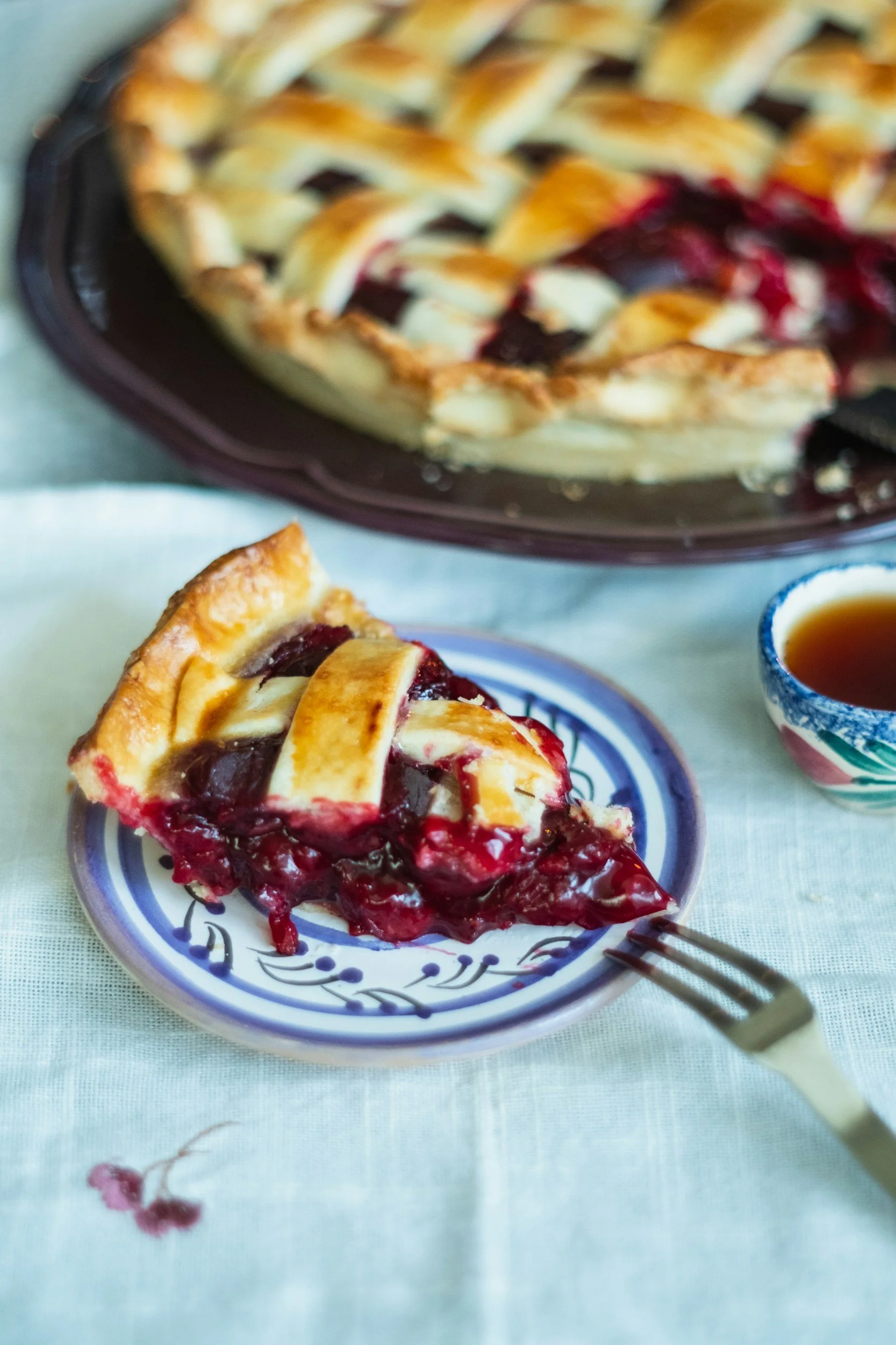 Slice of cherry berry pie with lattice crust on a small decorative blue and white plate, with a fork beside it, positioned on a white tablecloth next to a cup of tea, with the remaining pie in the background.