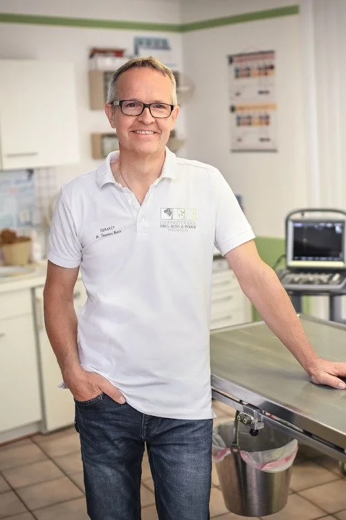 Ein Mann steht in einer Tierklinik, lächelnd, trägt eine weiße Klinikuniform mit Aufdruck, im Hintergrund sind medizinische Geräte und Poster sichtbar.