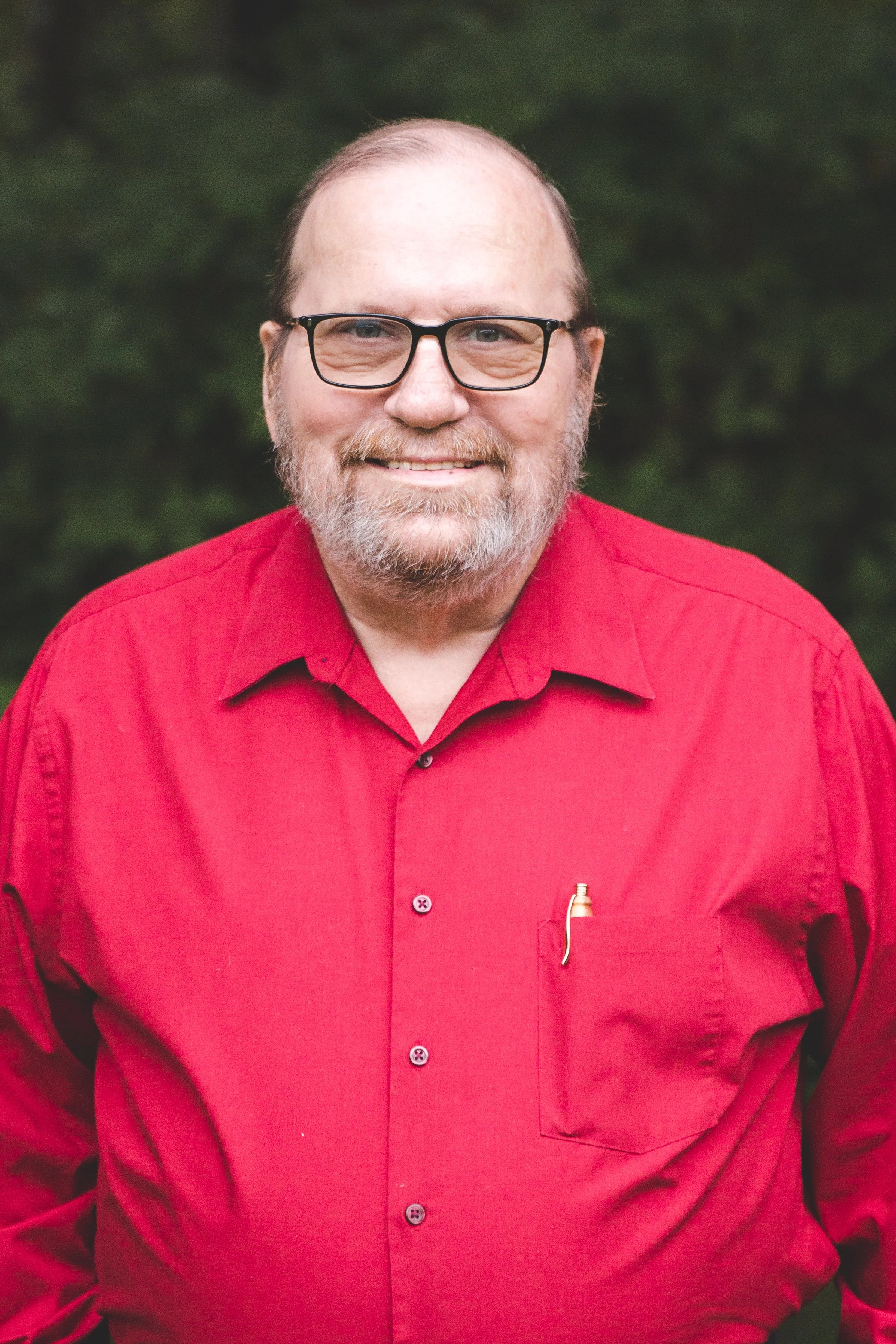 A smiling man with glasses, a beard, and a mustache, wearing a red shirt outdoors with a blurred green background.