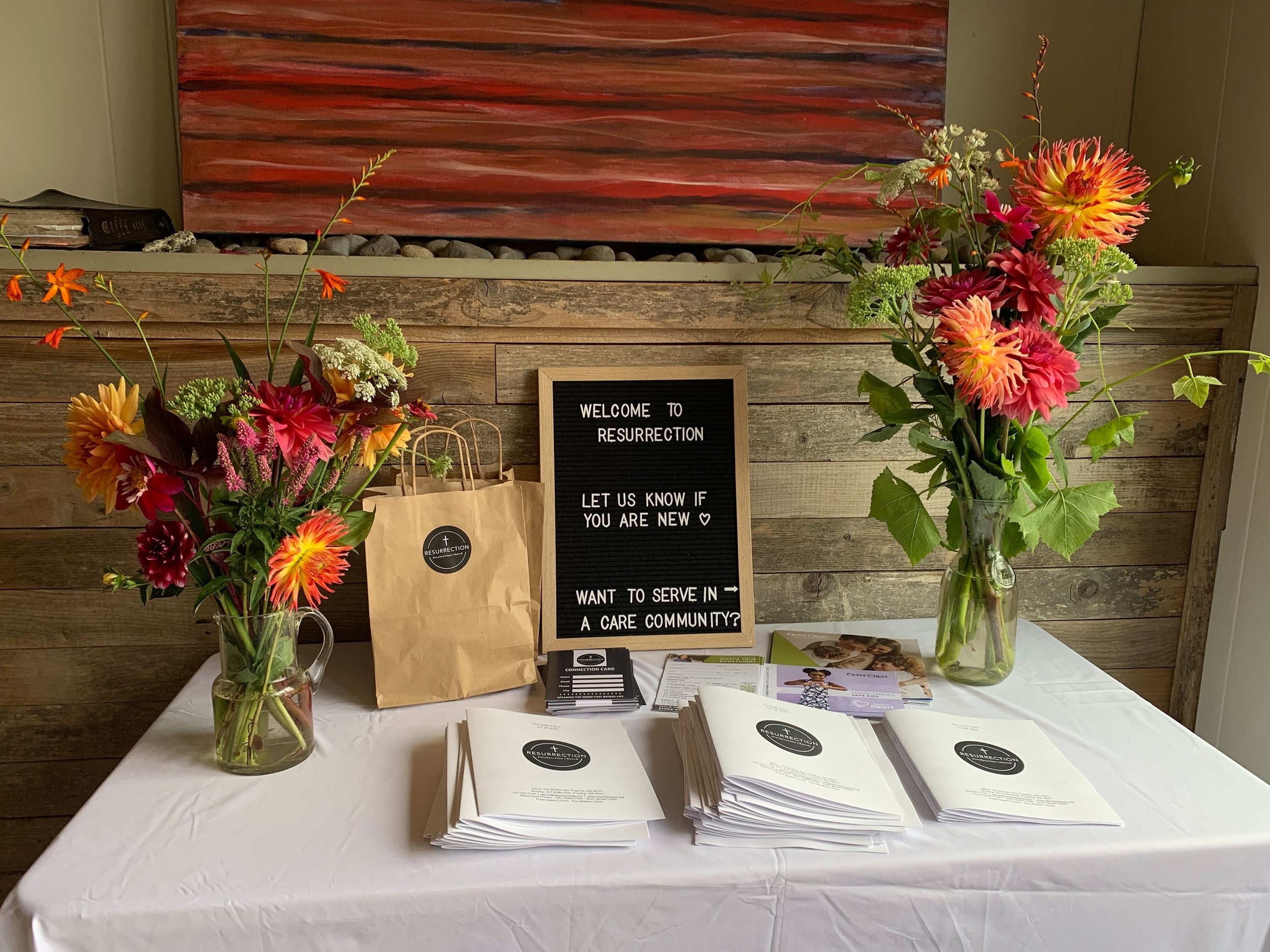 Table with informational flyers, brochures, and business cards, flanked by two large vases of colorful flowers, and a chalkboard sign with white text about offering a care community.