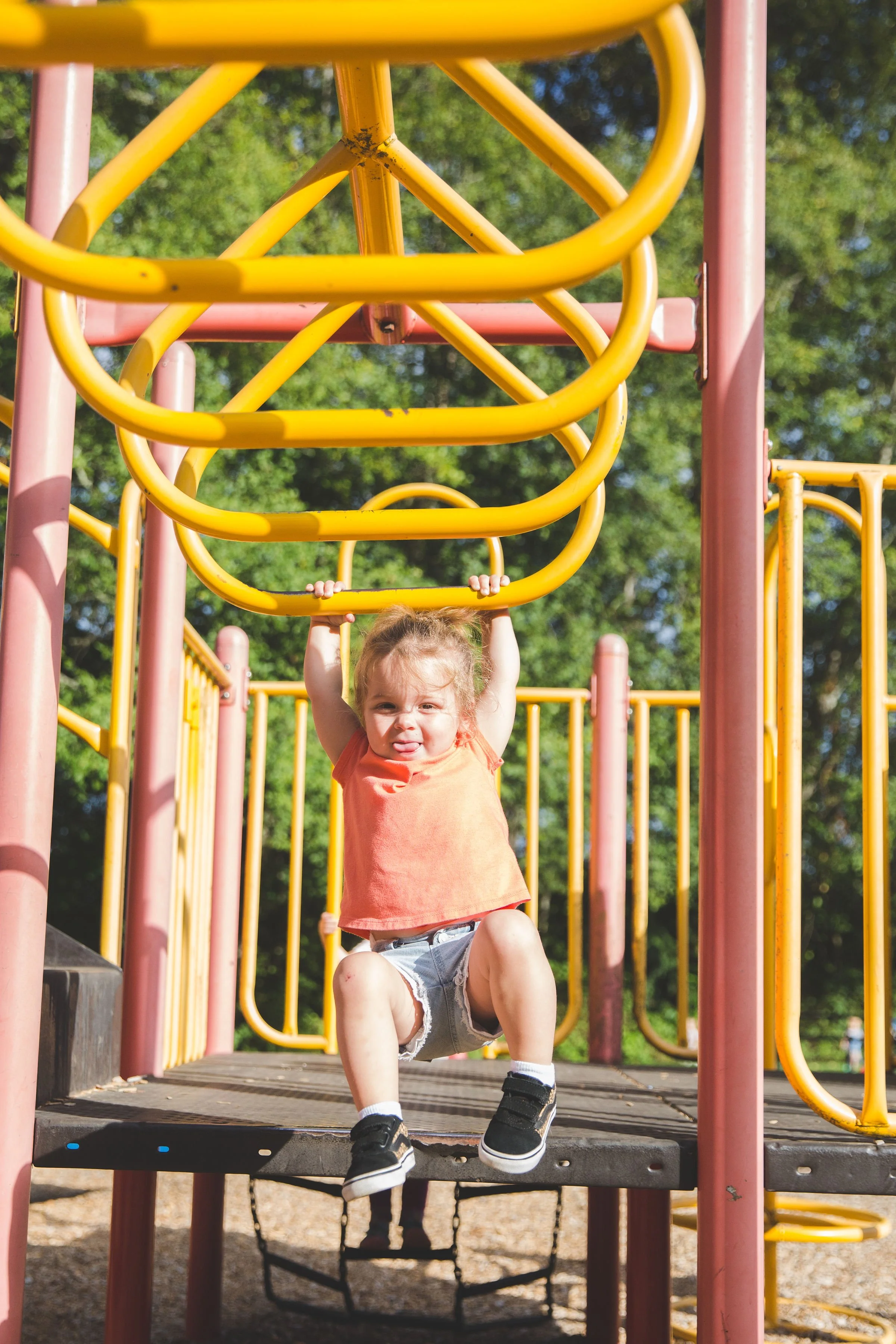 A young girl hanging from a yellow and pink jungle gym at a playground outdoors on a sunny day.