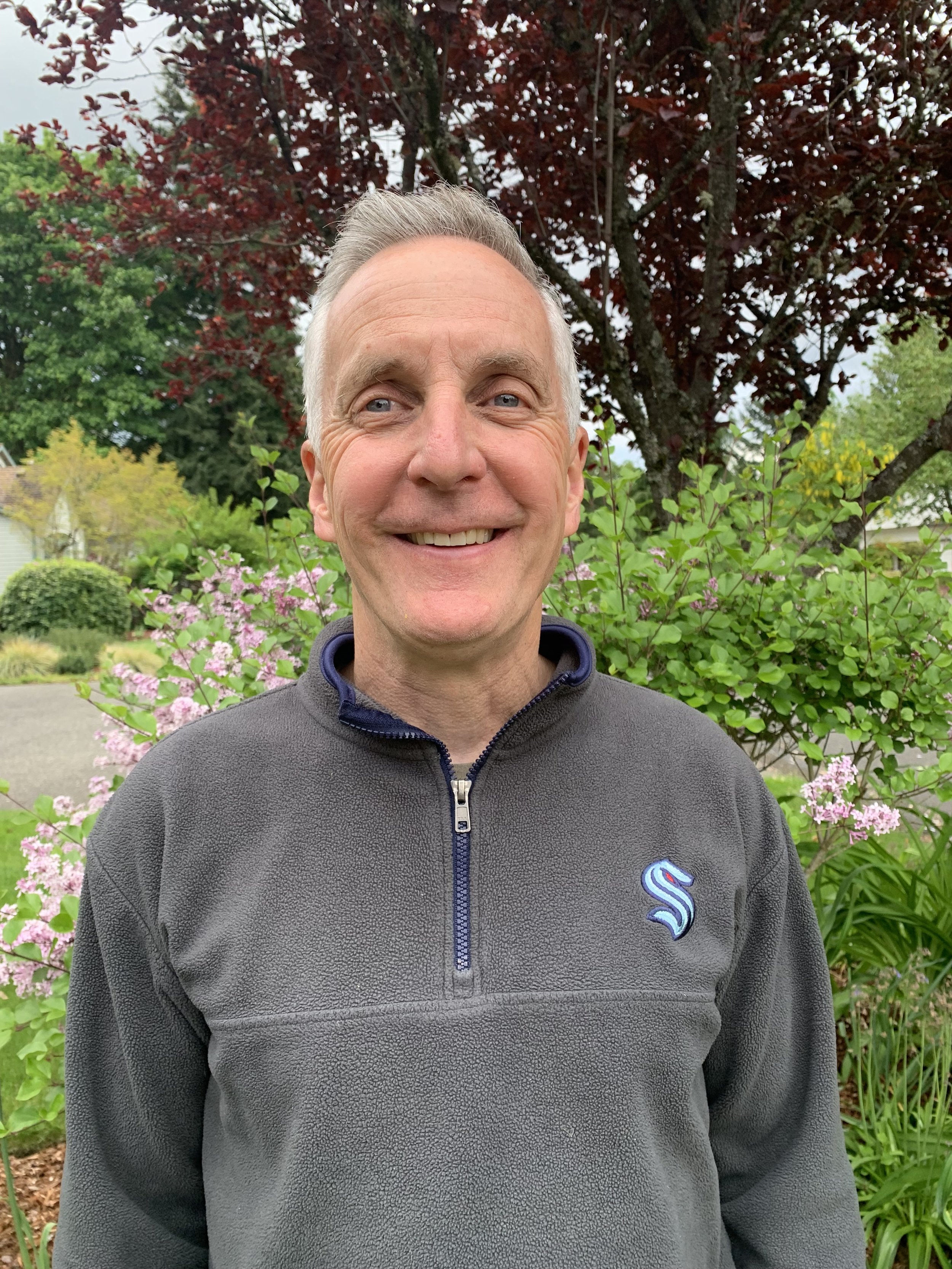 A smiling middle-aged man with short gray hair, wearing a gray fleece pullover with a zipper and a blue logo on the chest, standing outdoors in front of a garden with pink flowers, green bushes, and a dark red tree.