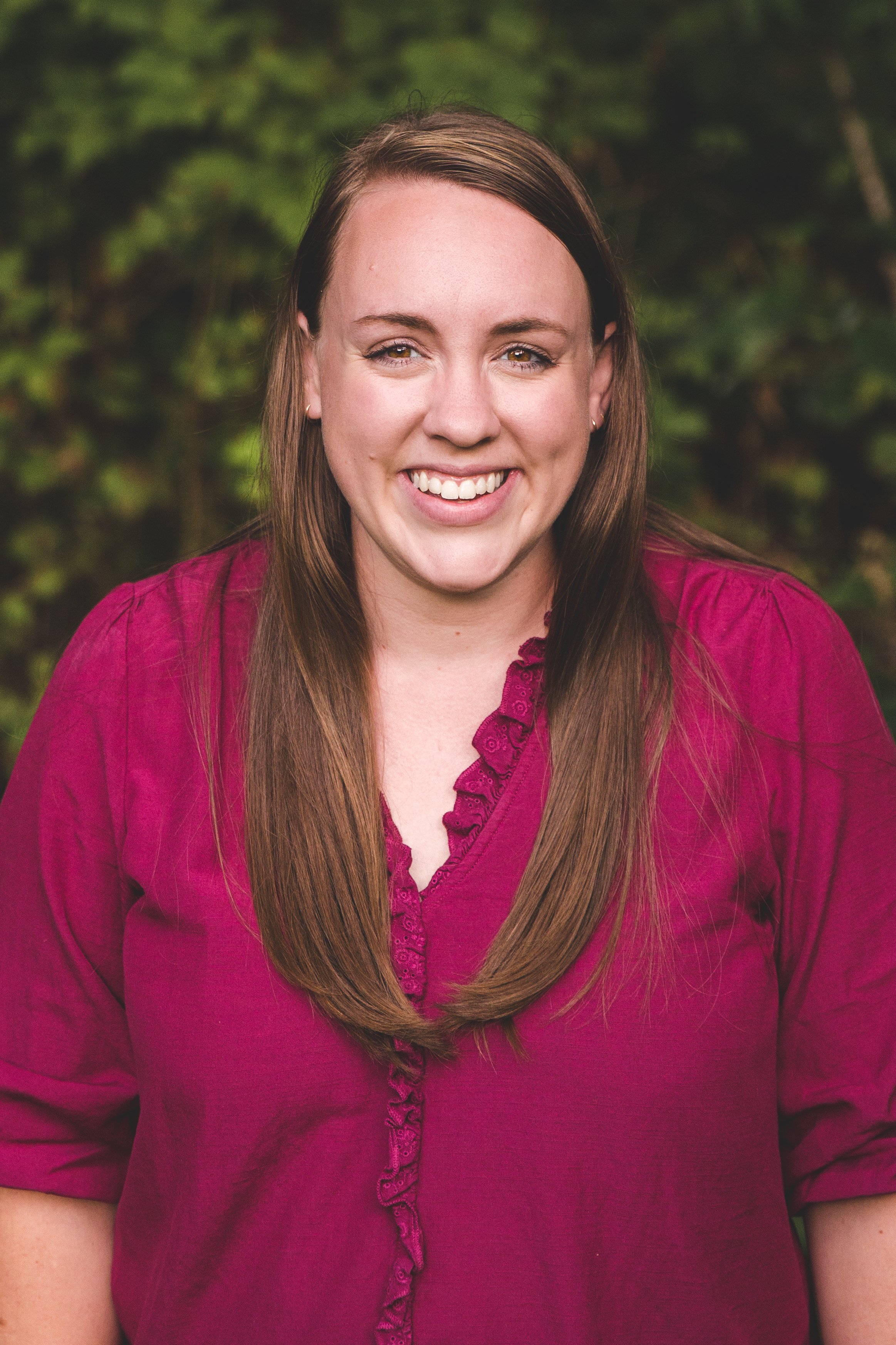 A woman with long brown hair smiling outdoors, wearing a burgundy top with ruffle details, with greenery in the background.