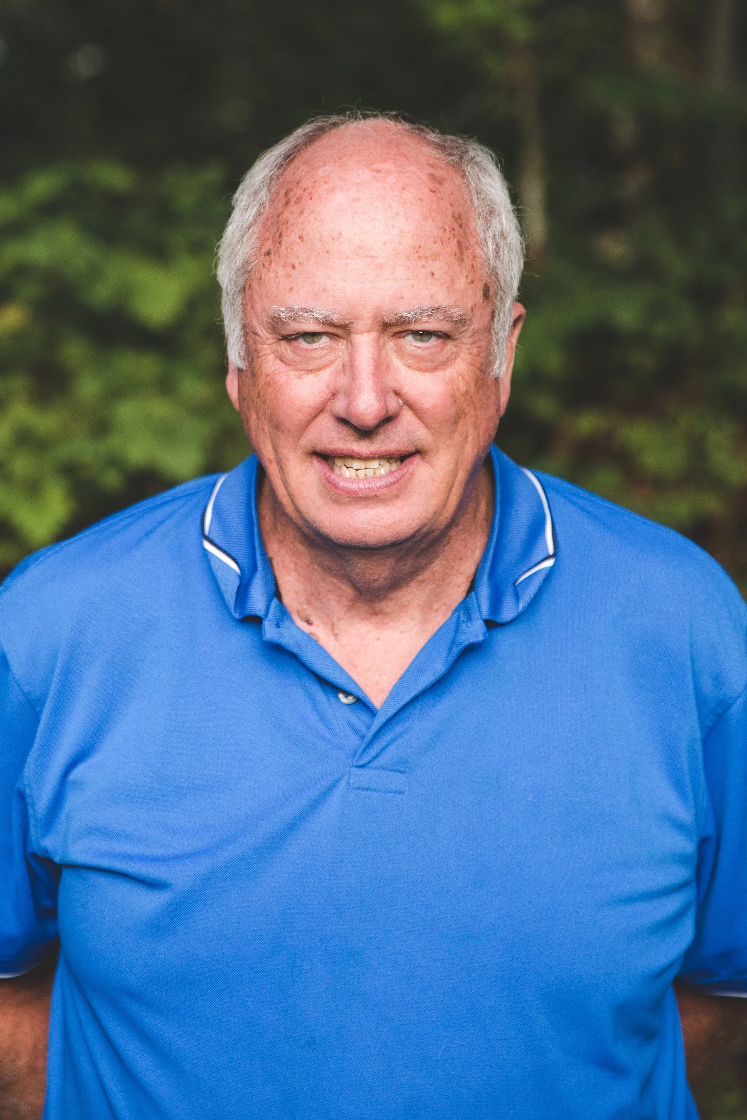 An elderly man with short gray hair and freckles, wearing a blue polo shirt, outdoors in a green natural setting.