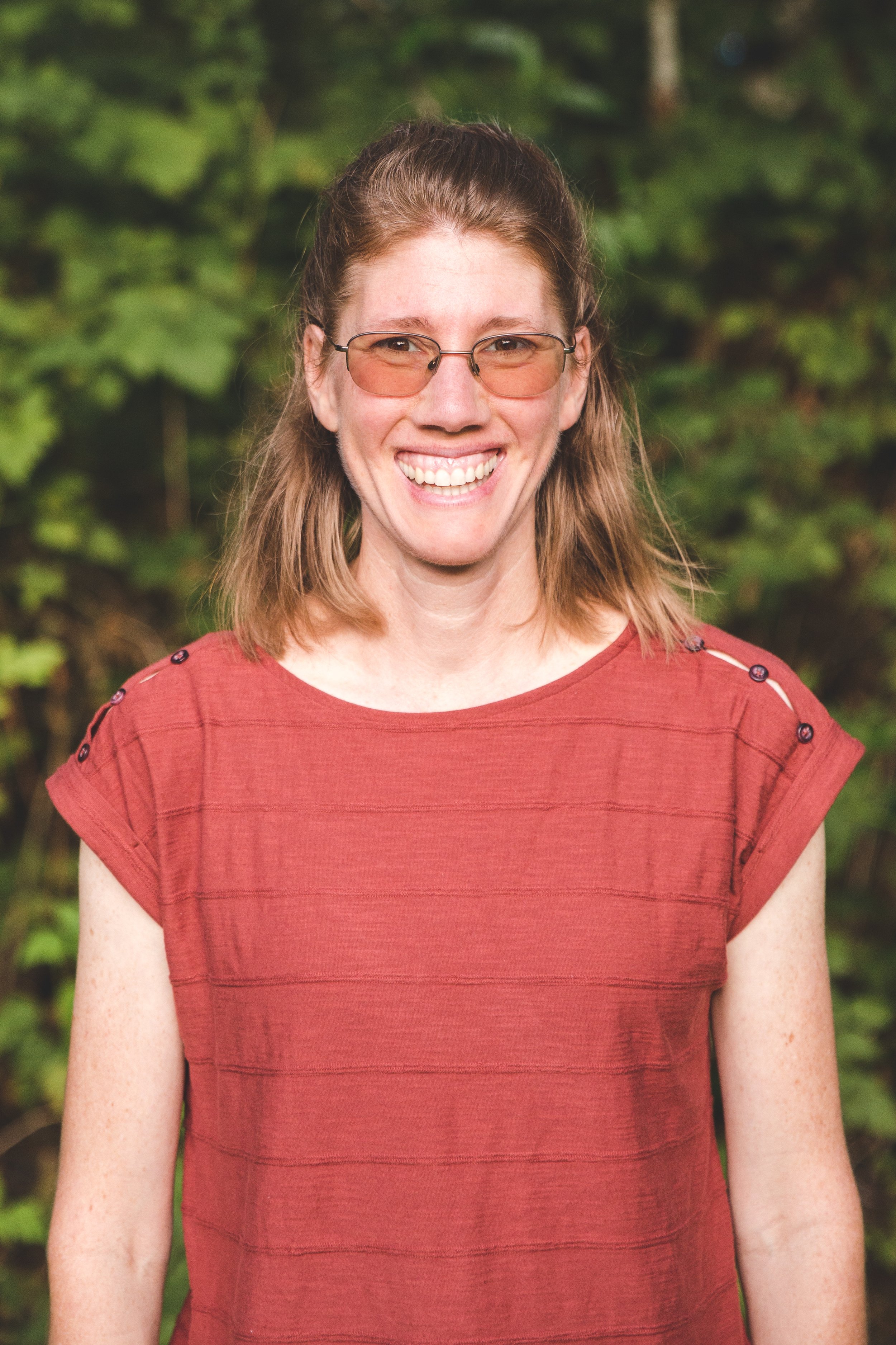 A smiling woman with shoulder-length light brown hair wearing glasses with tinted lenses and a red top with shoulder buttons, standing outdoors with green foliage in the background.
