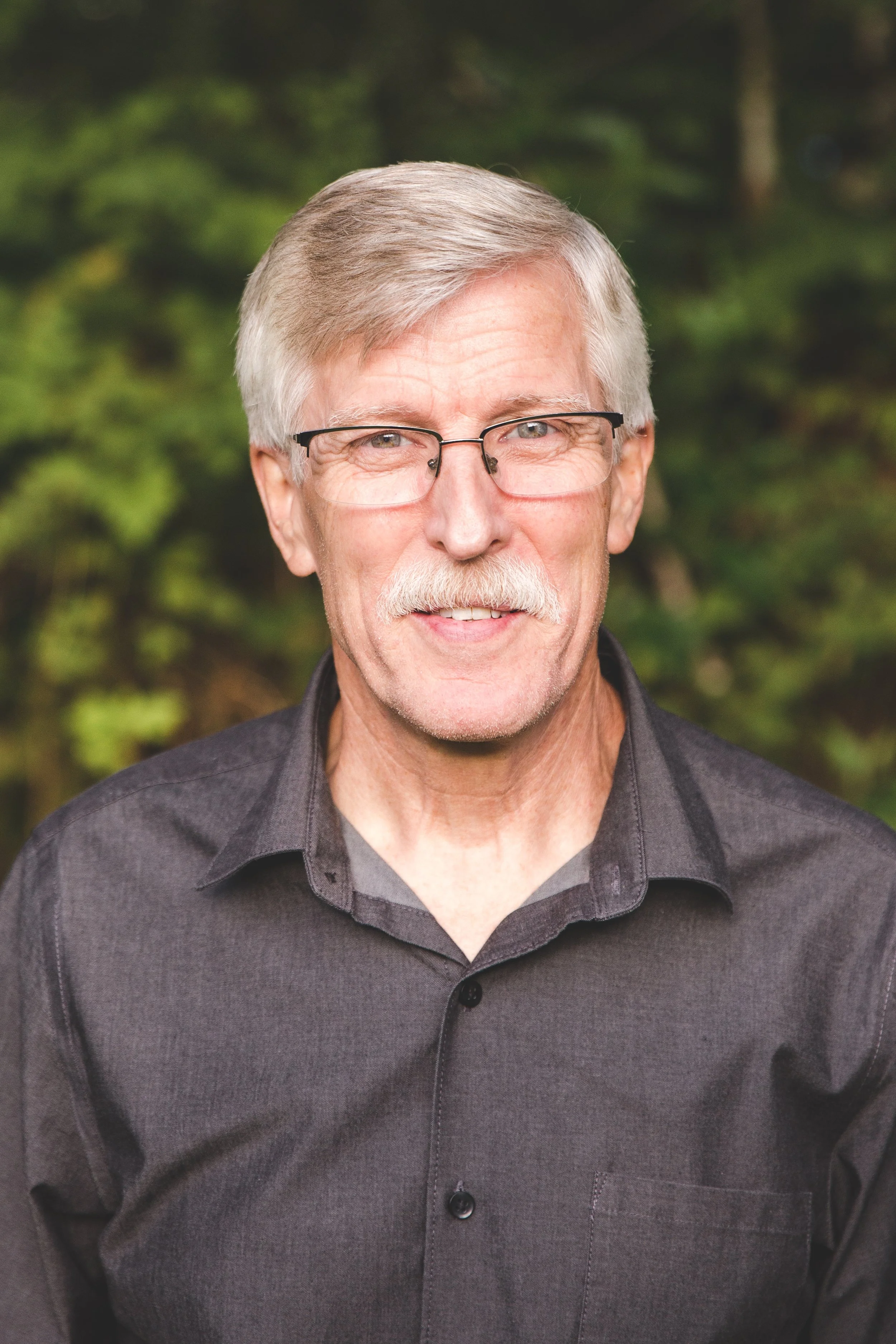 Close-up of an elderly man with gray hair and a mustache, wearing glasses and a dark gray shirt, outdoors with a blurred green forest background.